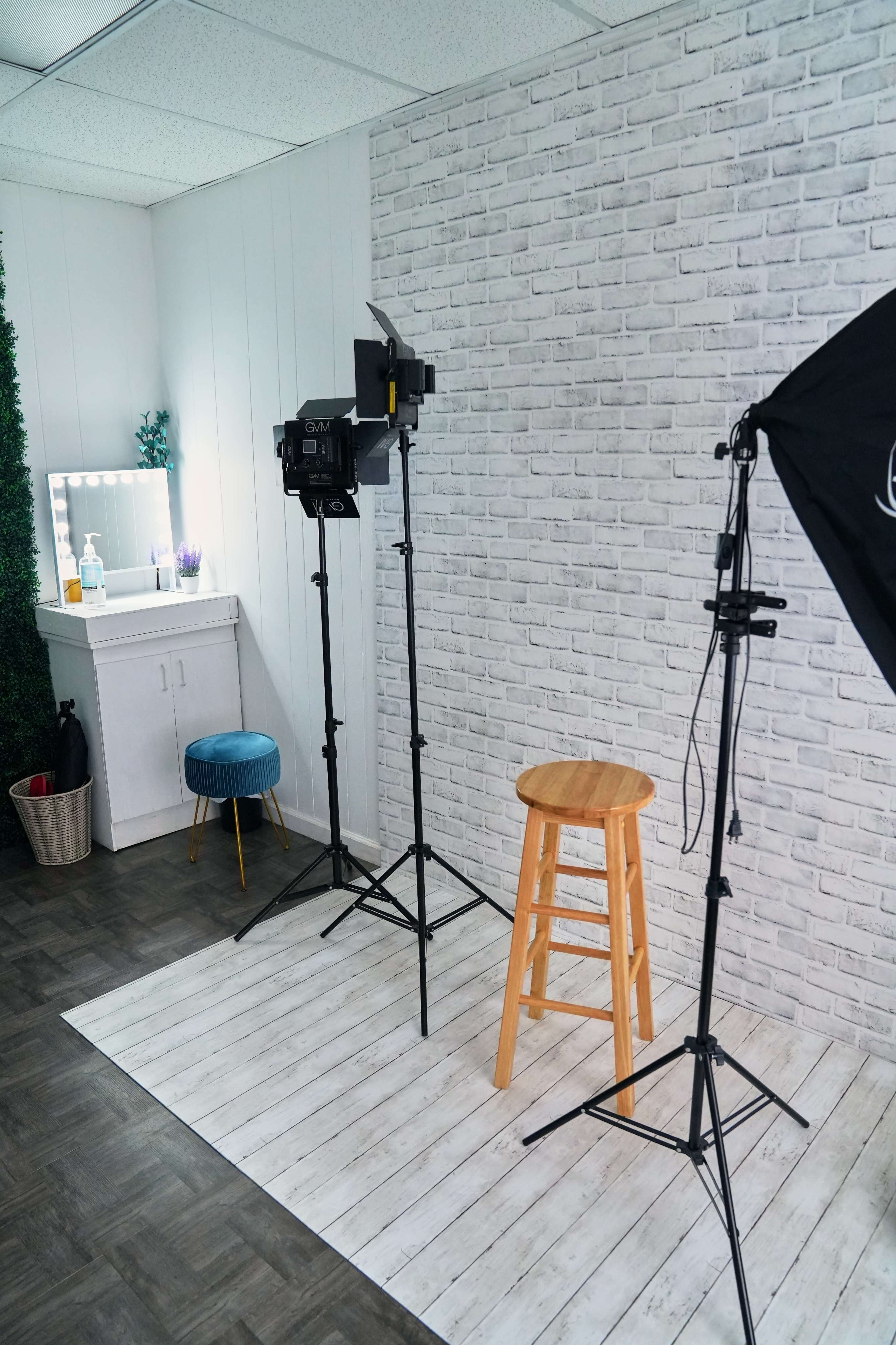The image shows a photography setup featuring a wooden stool on a light-colored floor, two softbox lights on tripods, and a mirror with a vanity area in the background.