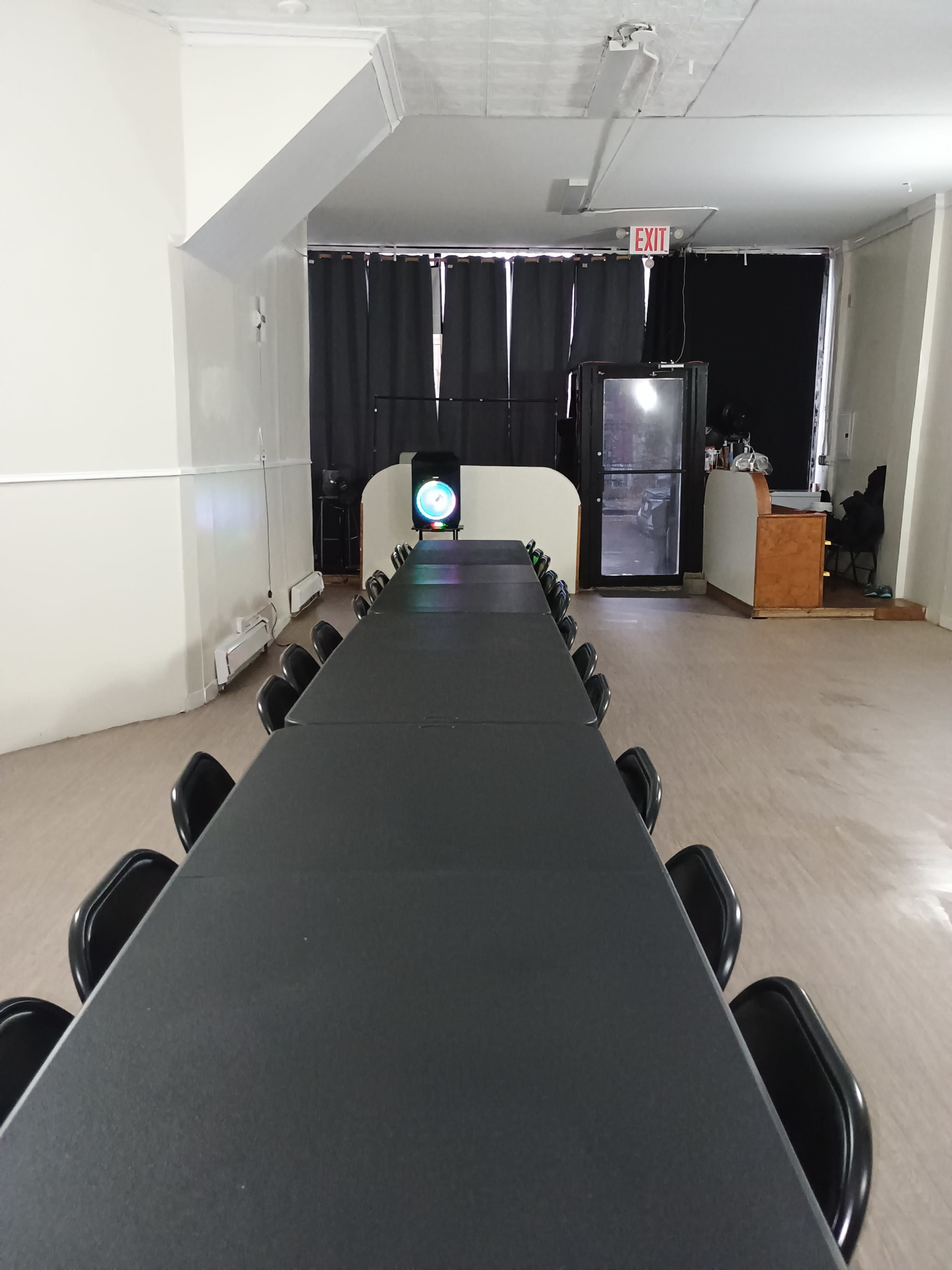 The image shows a long rectangular table with black chairs arranged on either side, set in a room with dark curtains and a visible exit sign.