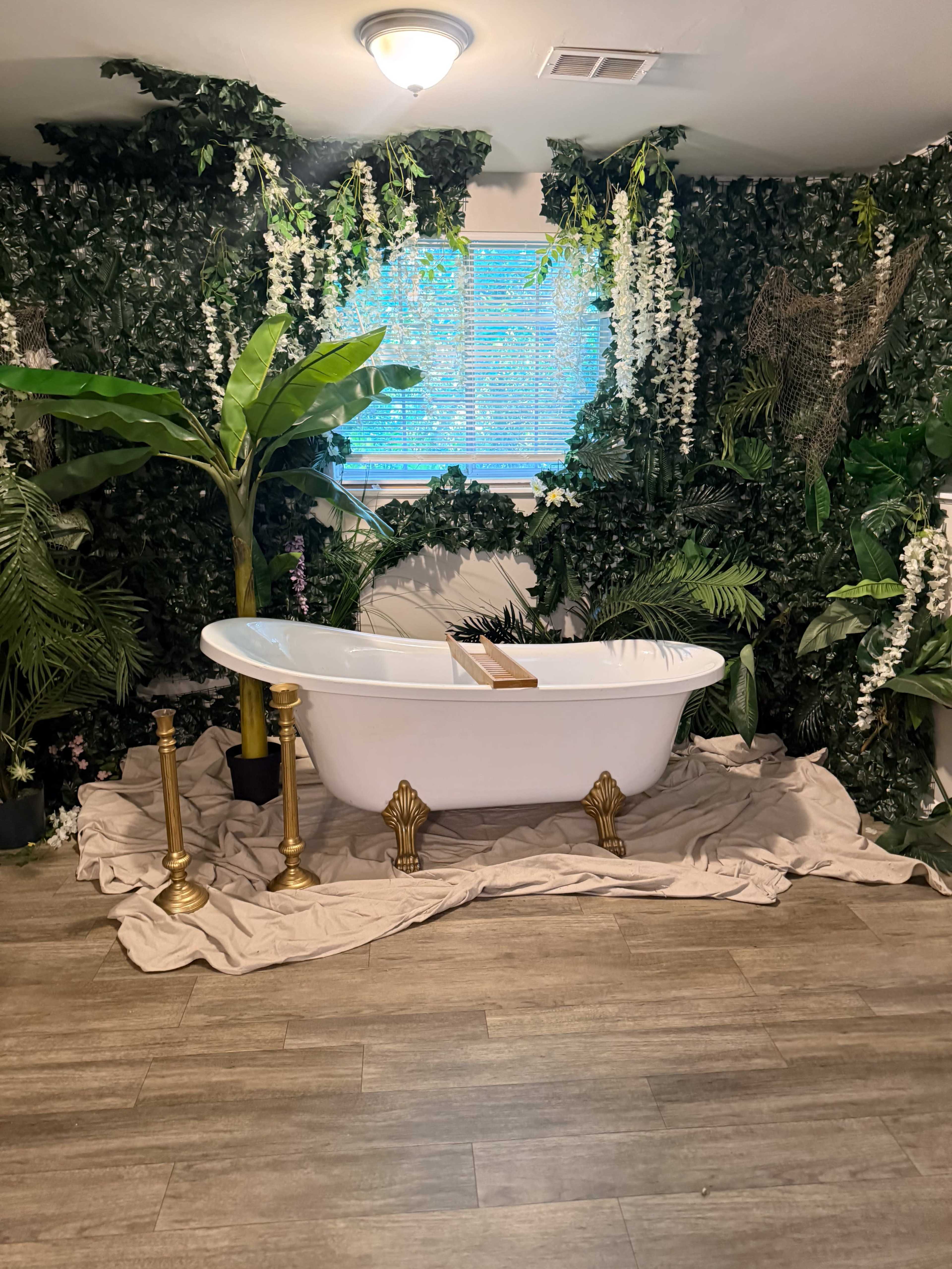 The image shows a freestanding white bathtub placed on a beige mat, surrounded by lush green plants and decorative foliage against a wall.