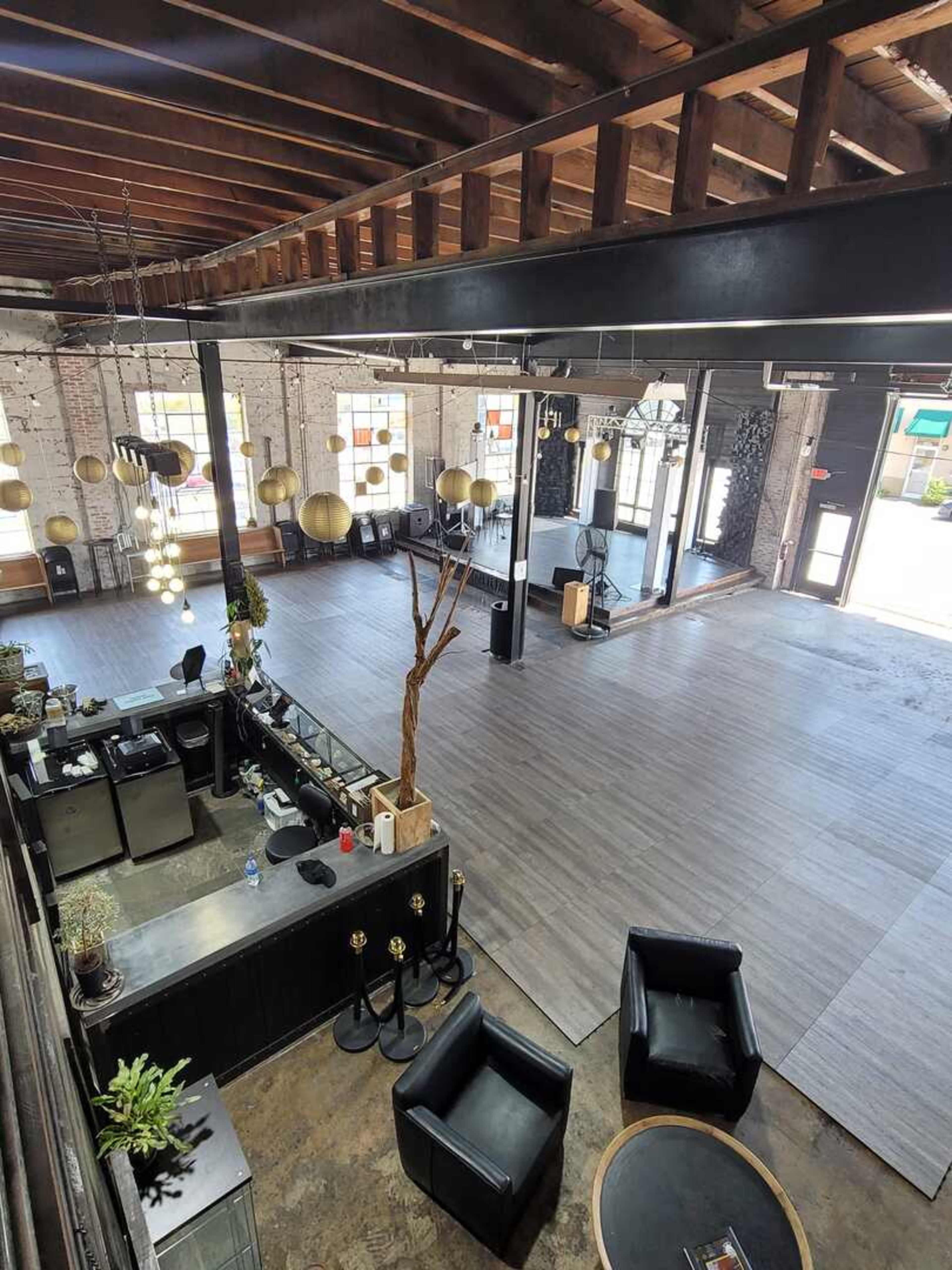 The image shows an open, expansive interior space with high ceilings, wooden beams, and a reception area on the left, featuring modern furnishings and decorative lighting.