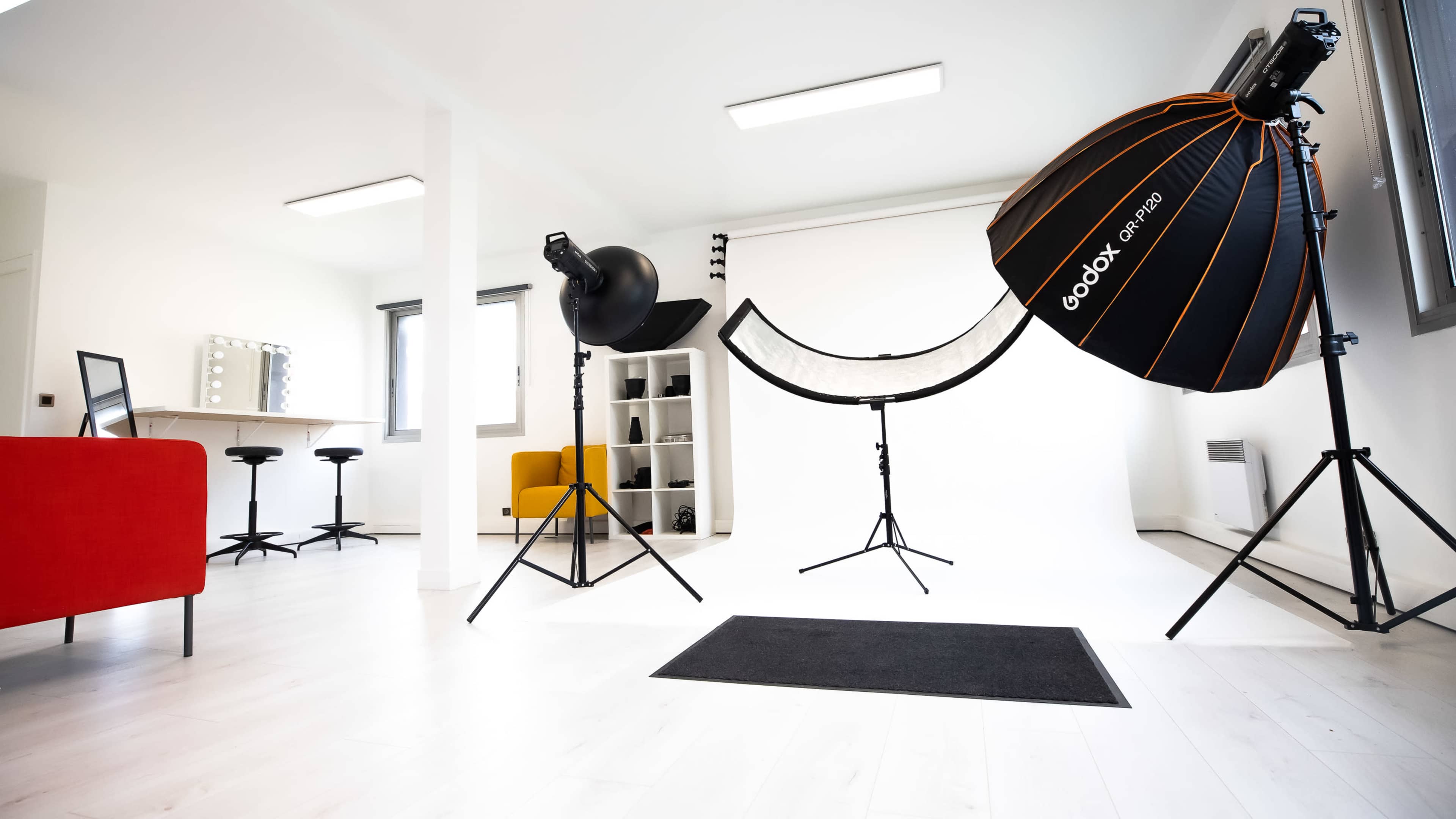 A bright photography studio is set up with lighting equipment, a backdrop, and various furniture pieces, including a red sofa and bar stools.