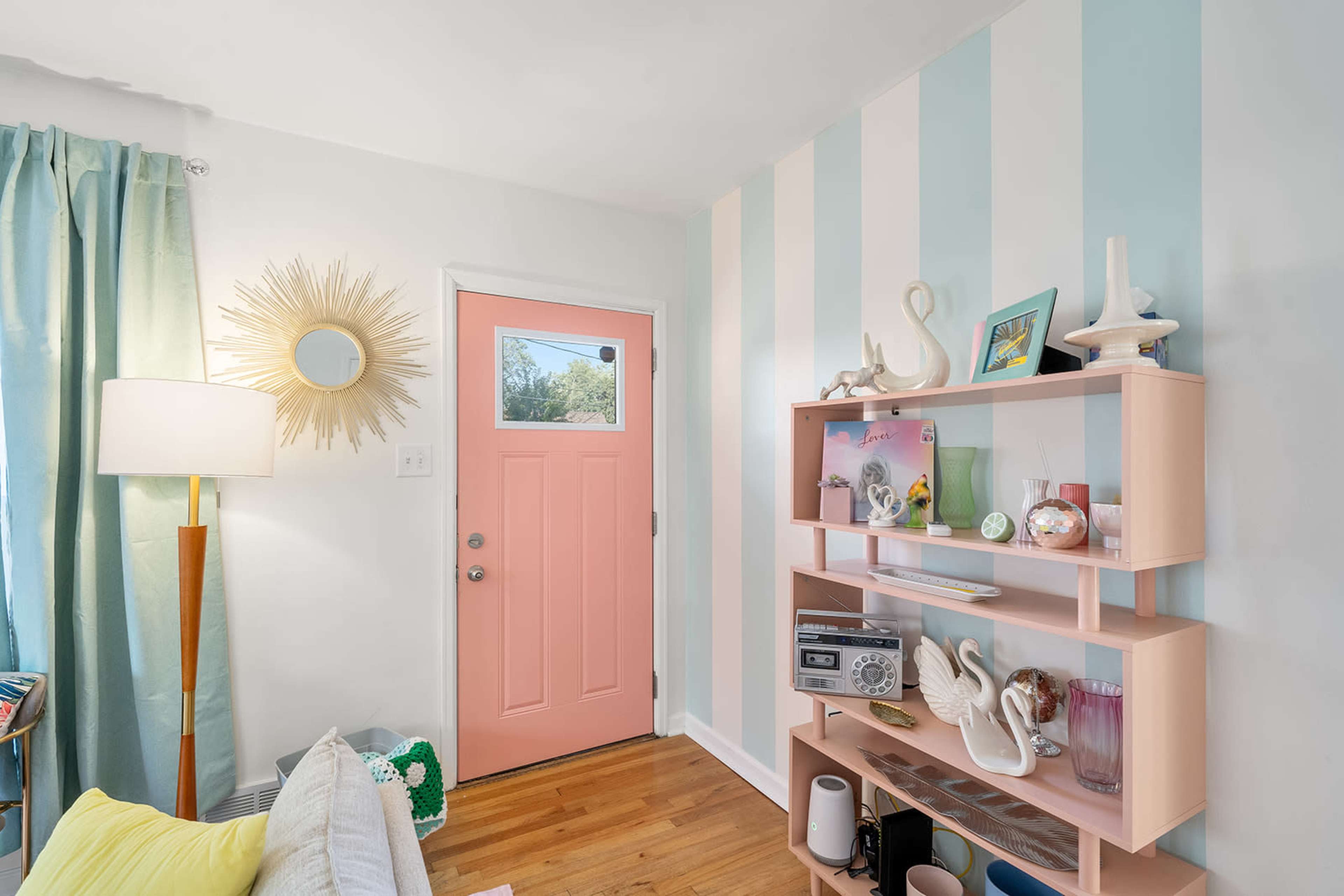 A brightly lit corner of a living room featuring a pastel pink door, striped blue and white walls, a wooden floor, a shelf with decorative items, and a lamp beside a window with light teal curtains.