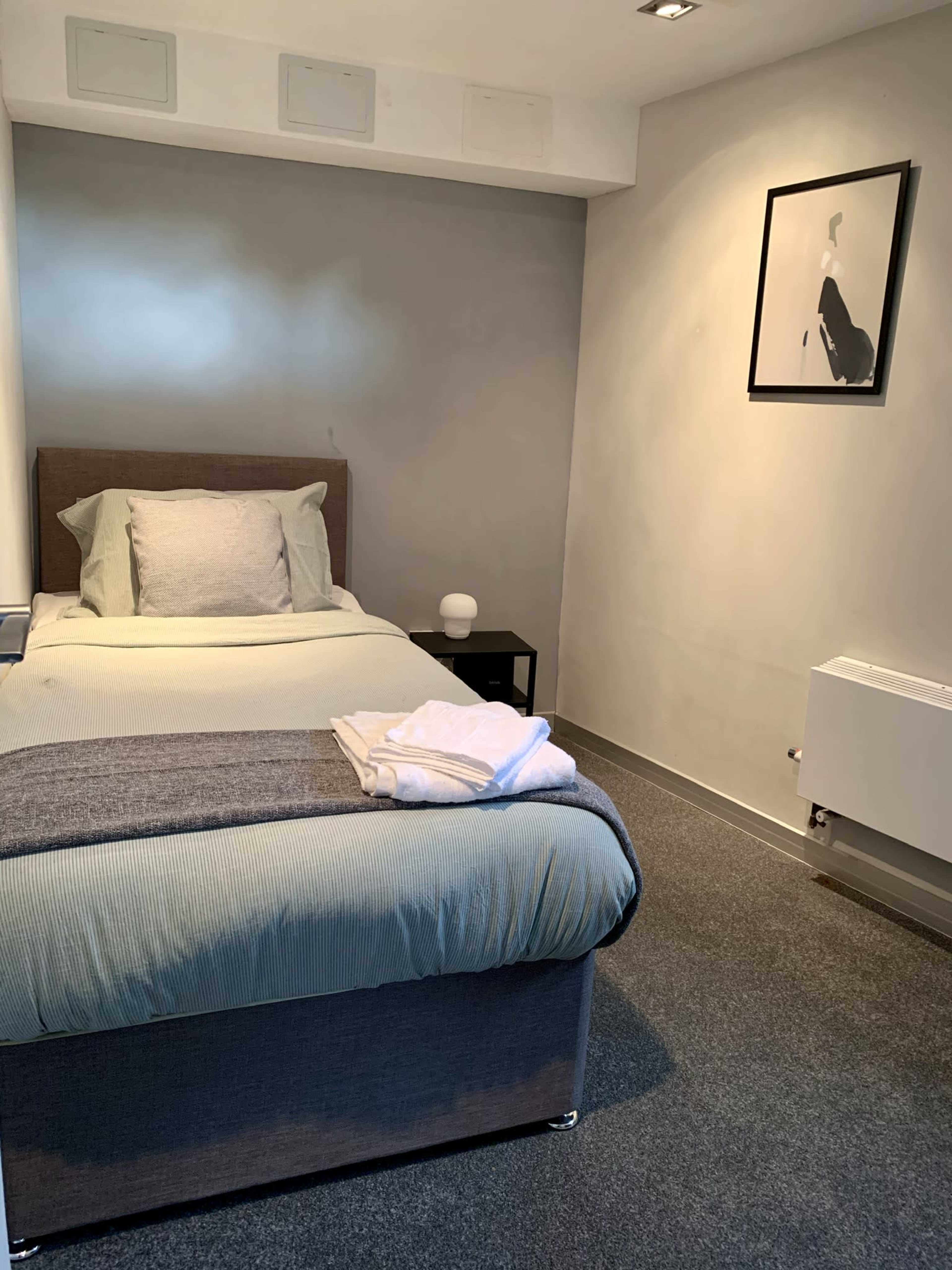A compact bedroom featuring a single bed with a gray and white color scheme, a bedside table with a lamp, and a framed artwork on the wall.