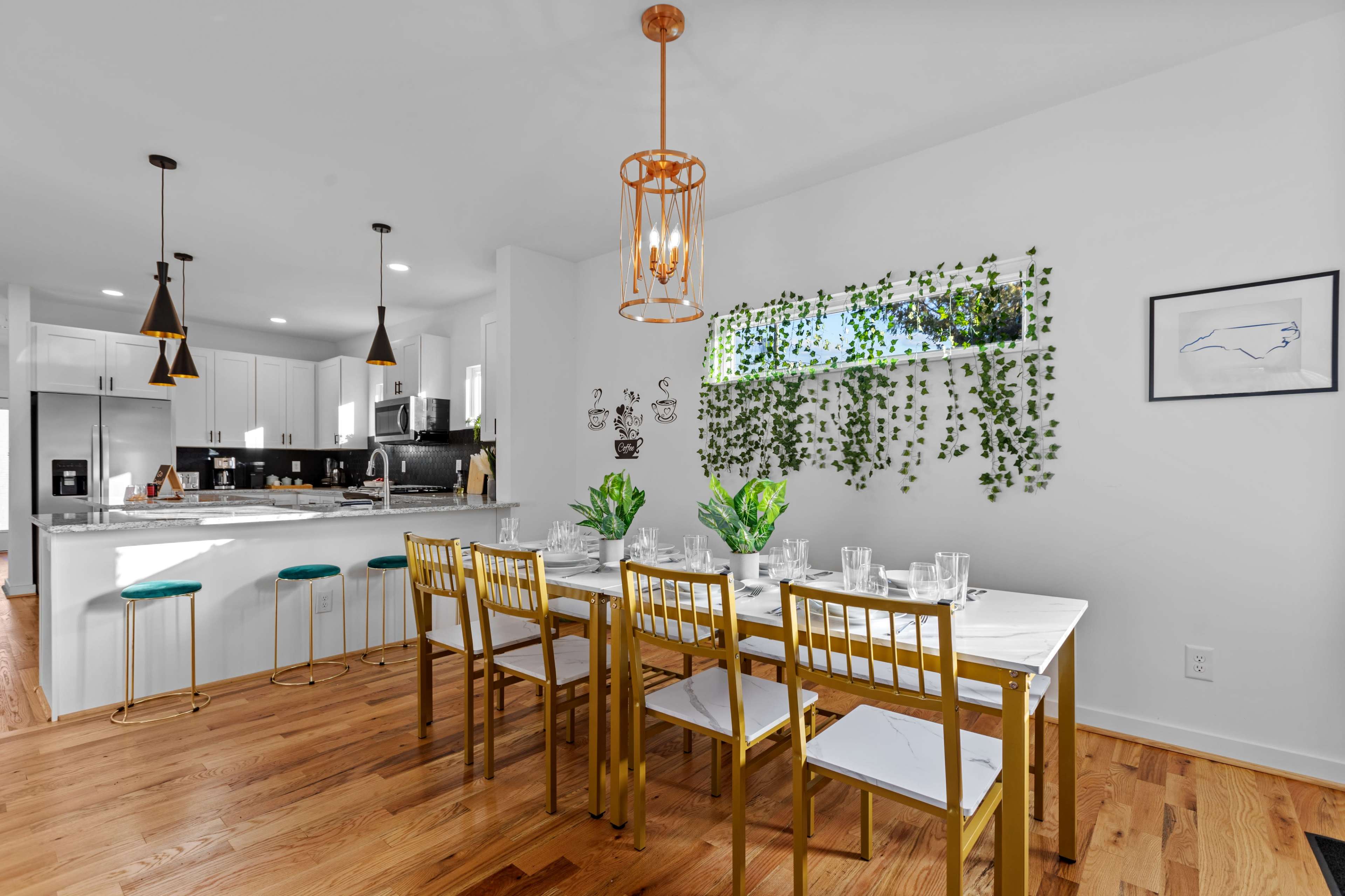 A modern dining area features a long table with gold chairs, surrounded by greenery and illuminated by pendant lights.