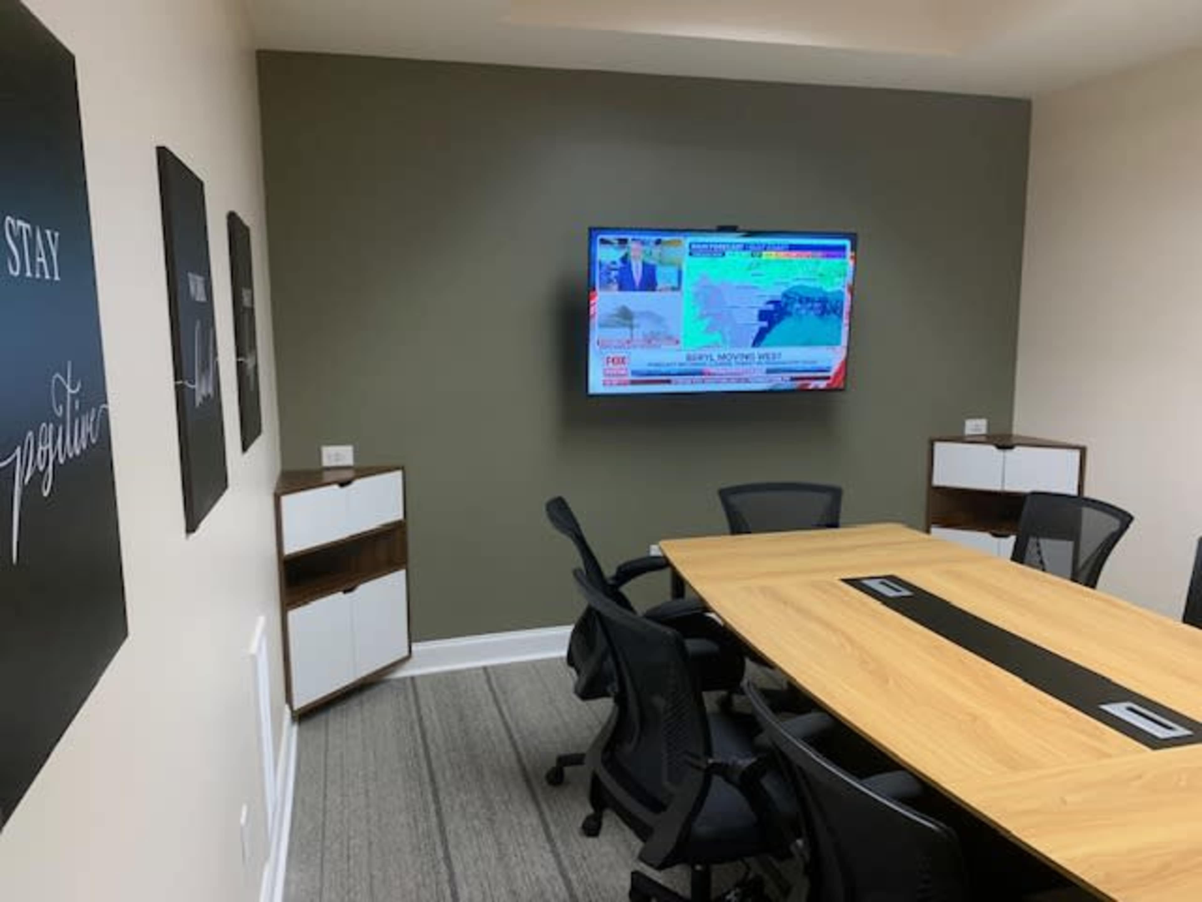 A conference room is equipped with a large television displaying news content, surrounded by a wooden table and black chairs.