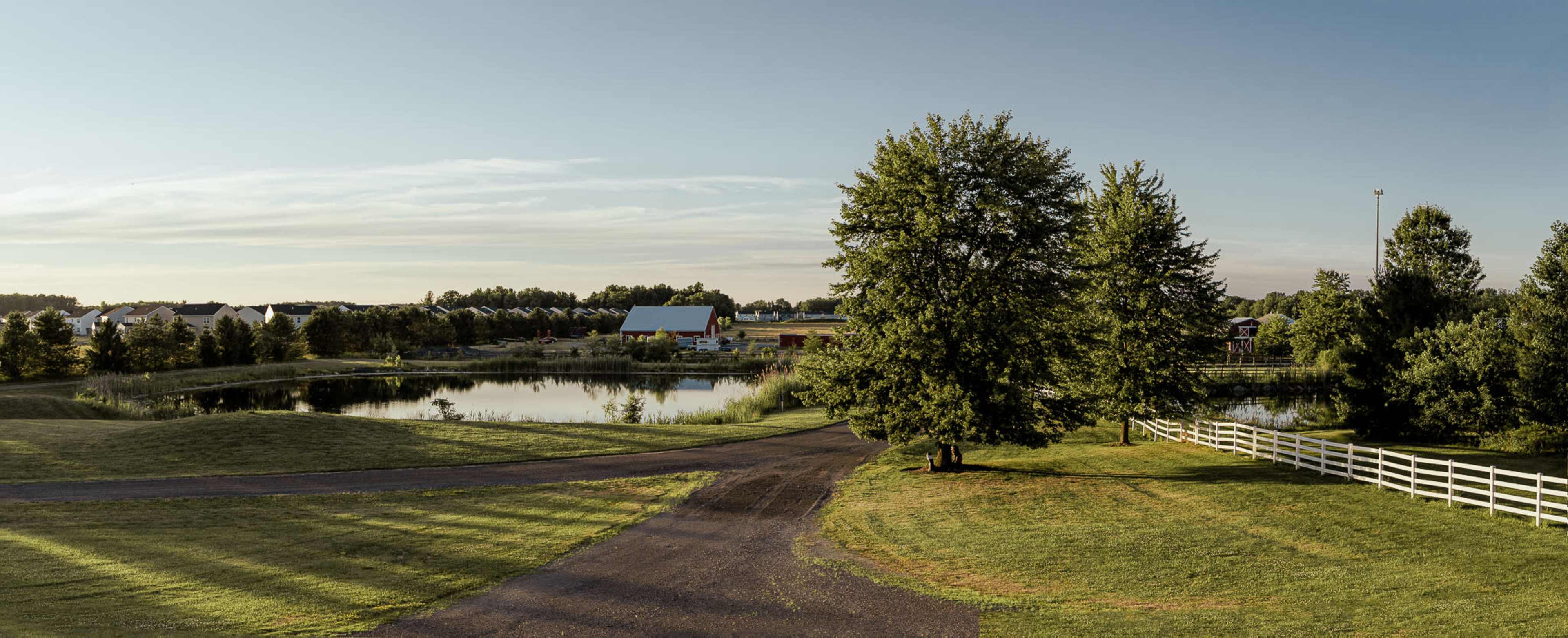 A winding path leads through a grassy area beside a pond, with trees and residential structures in the background.