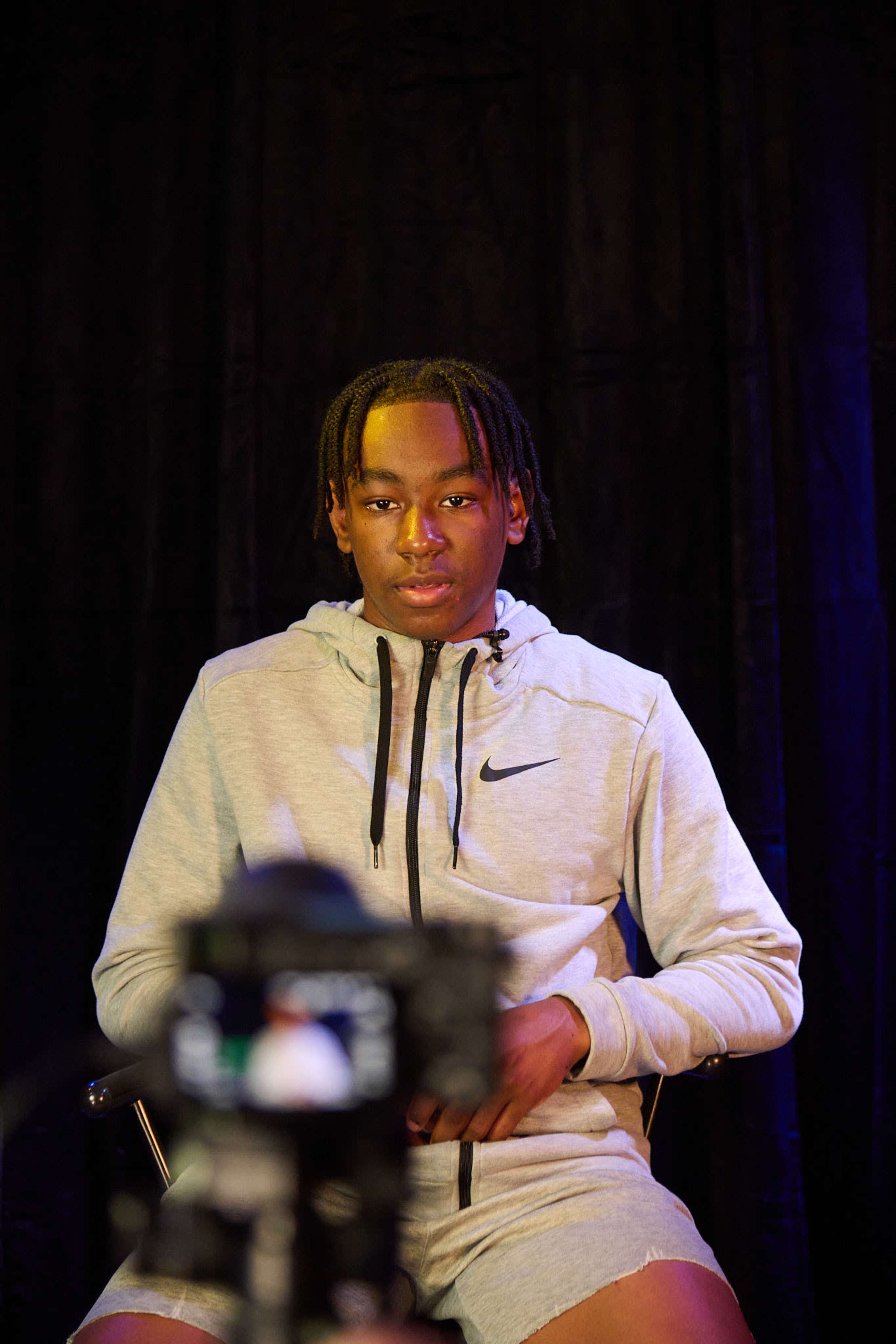 A young man in a gray Nike hoodie sits in front of a camera with a black backdrop.
