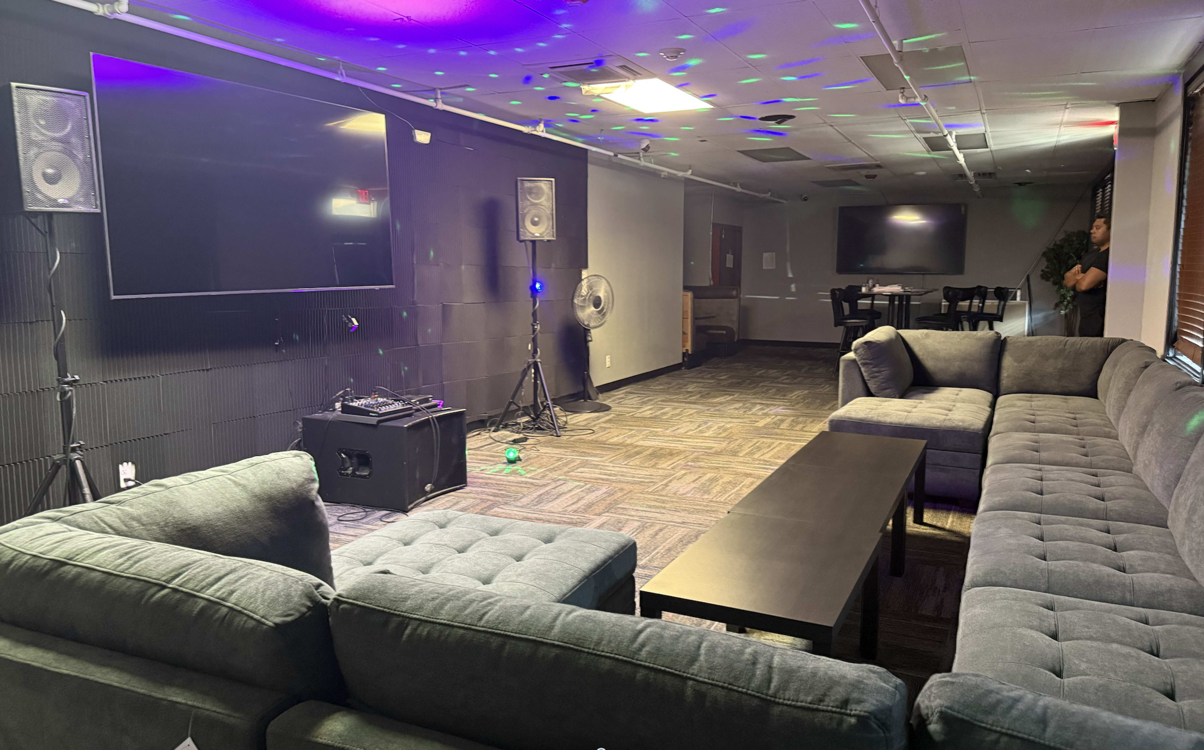 The image shows a lounge area with gray sofas, a black coffee table, speakers, and a large screen, illuminated by colored lights on the ceiling.