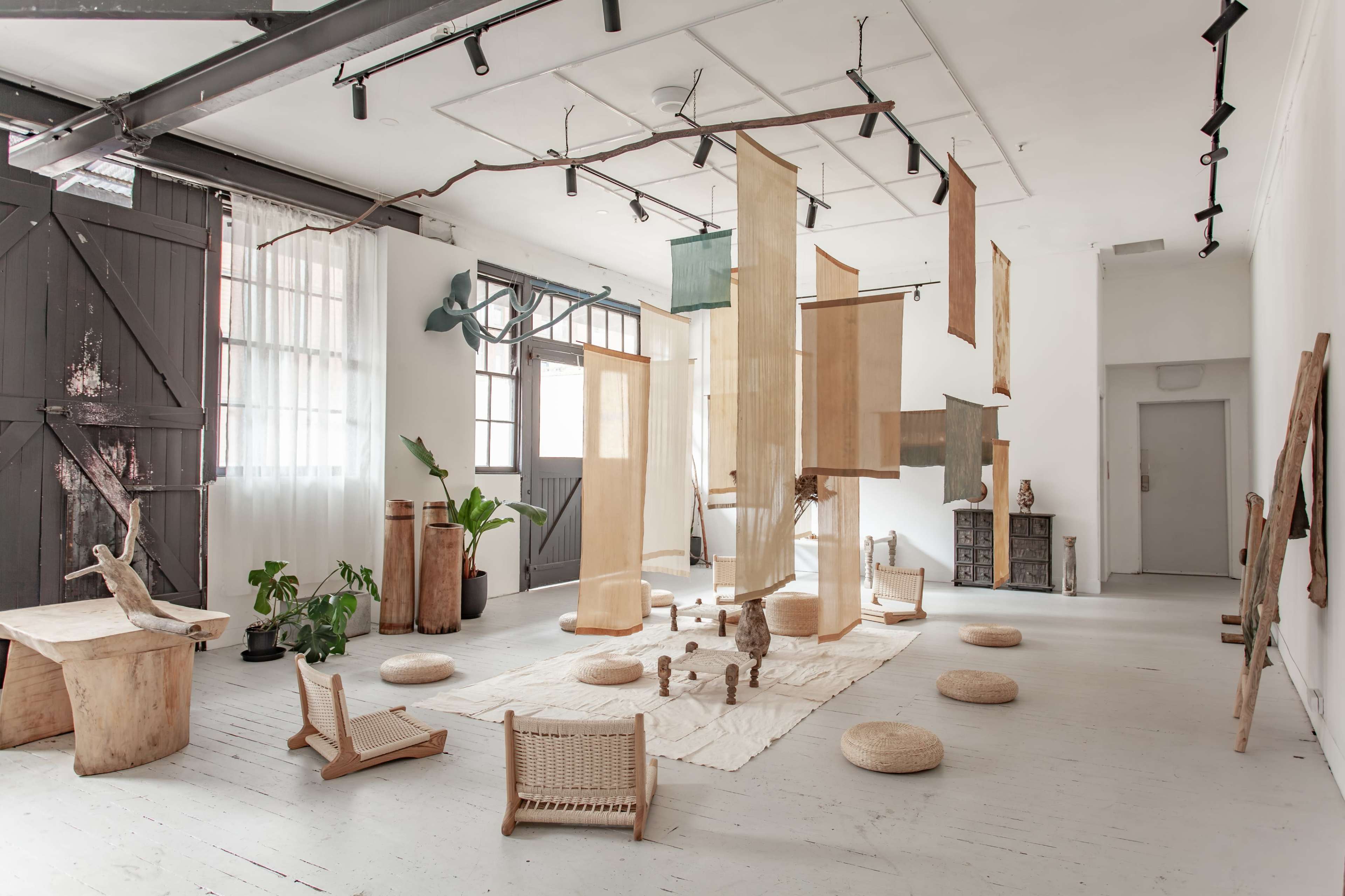 The image shows an art studio featuring various hanging fabric panels, wooden sculptures, and minimalist furniture arranged around a central area with light-colored flooring.