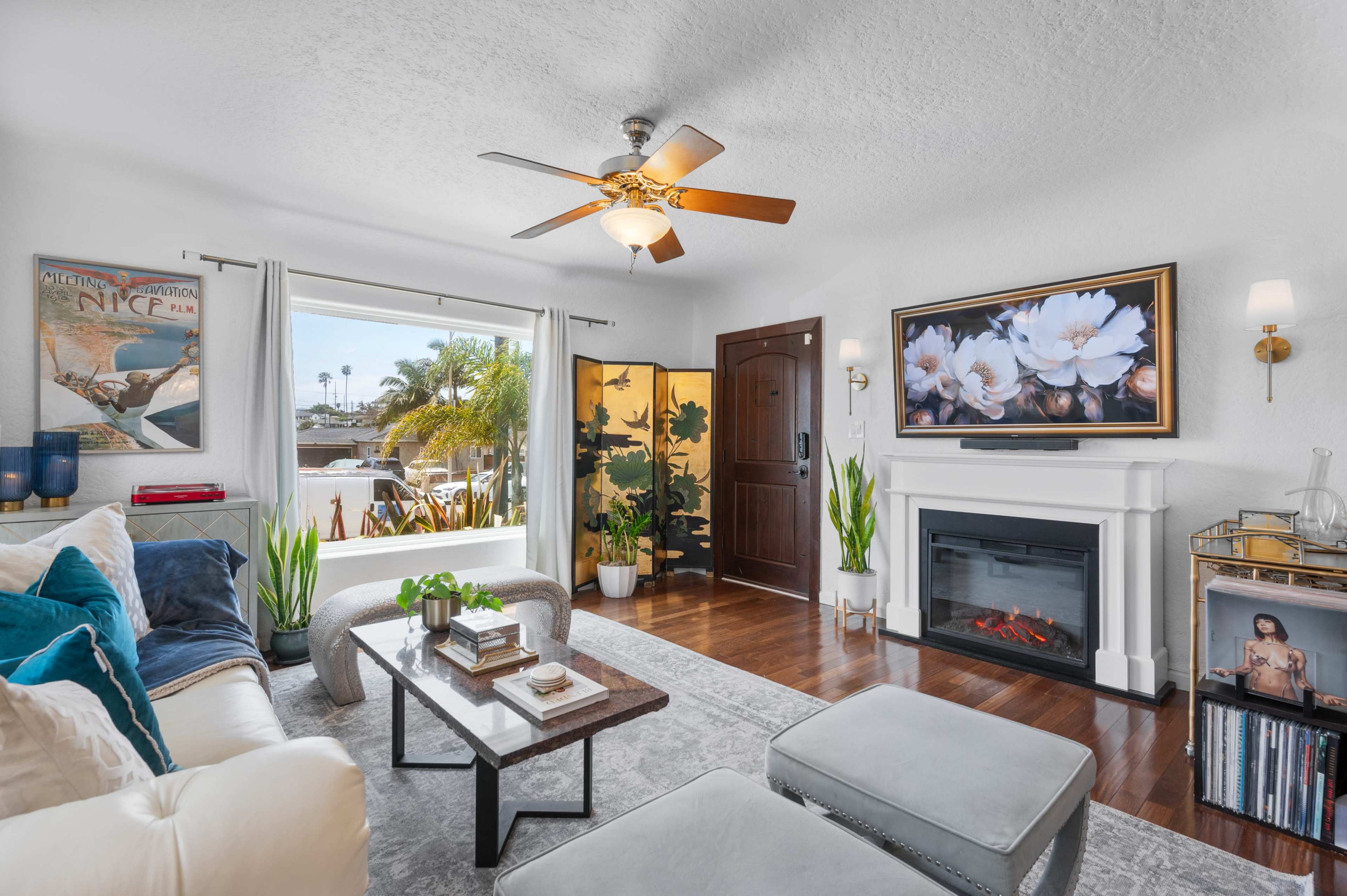 The living room features a sofa, a coffee table, and a decorative fireplace, with large windows showcasing a view of palm trees outside.