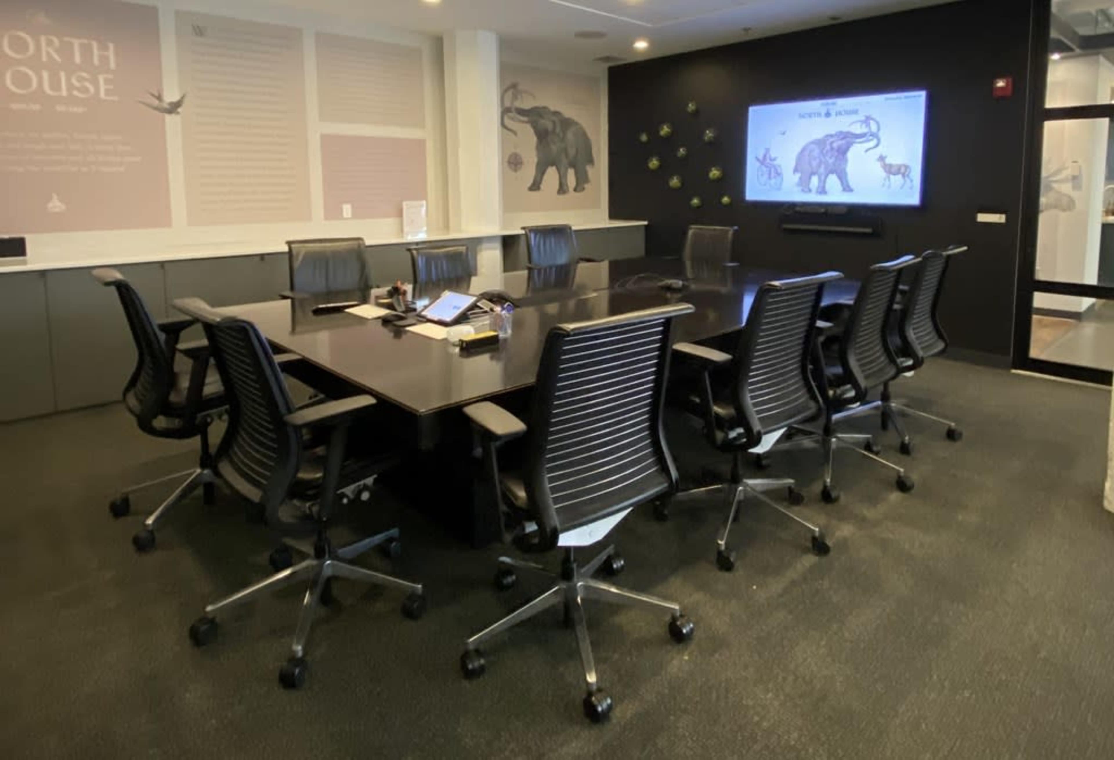 A conference room features a large black table surrounded by eight swivel chairs, with a screen displaying an elephant and walls adorned with art.