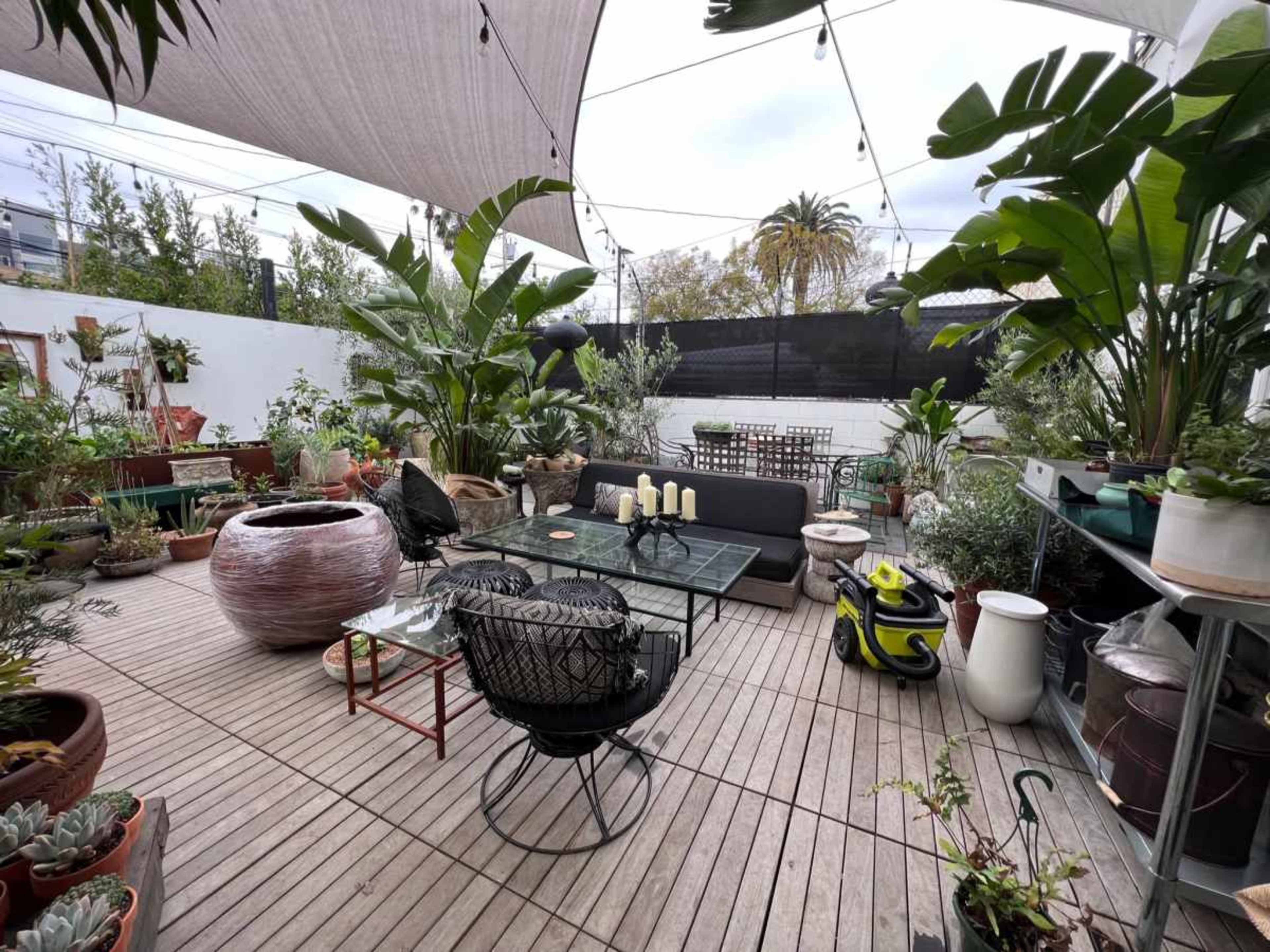 The image depicts a landscaped outdoor space featuring various potted plants, seating arrangements, and a wooden deck with a glass table.