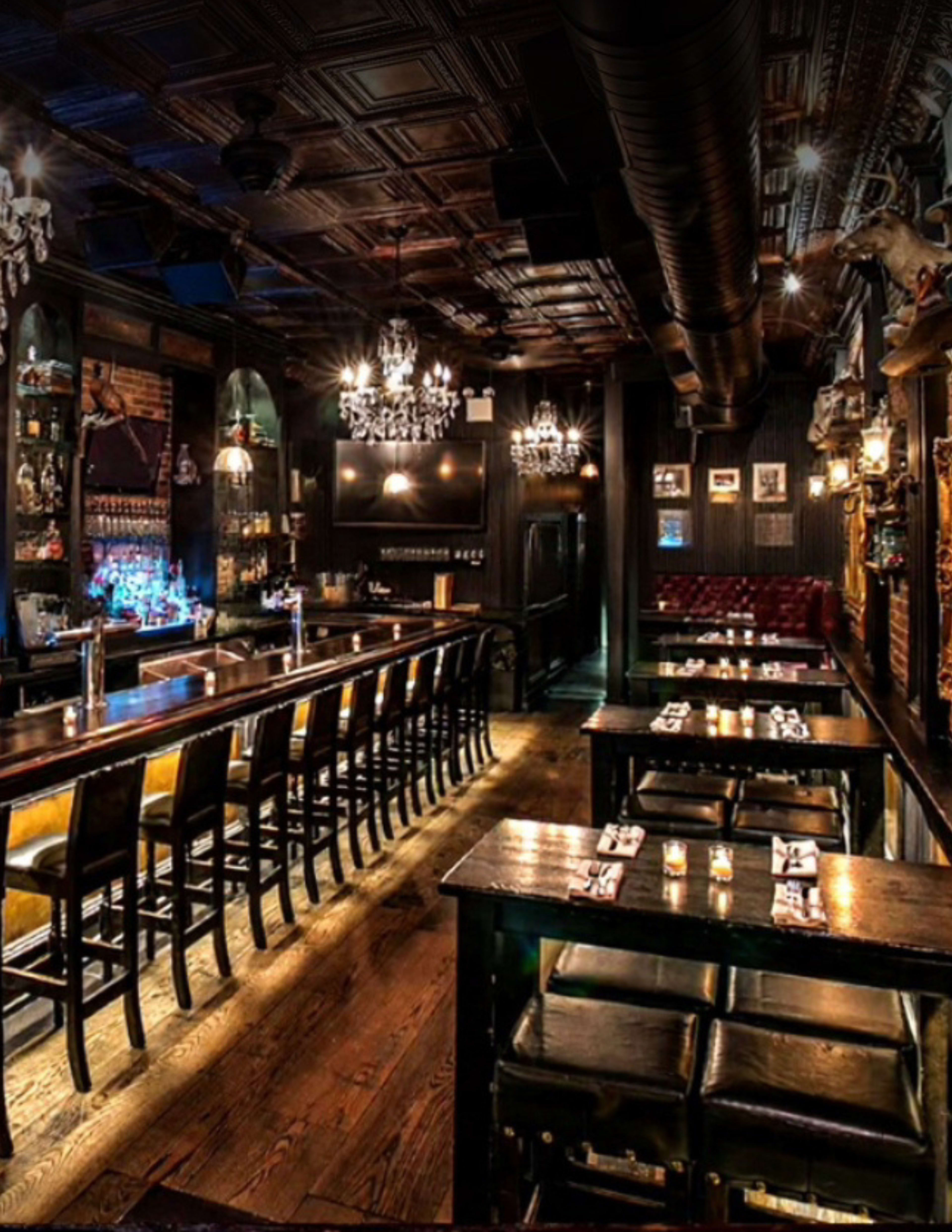 The image shows the interior of a bar with a long wooden counter, high stools, and several tables arranged in a dimly lit atmosphere adorned with chandeliers and dark wood paneling.