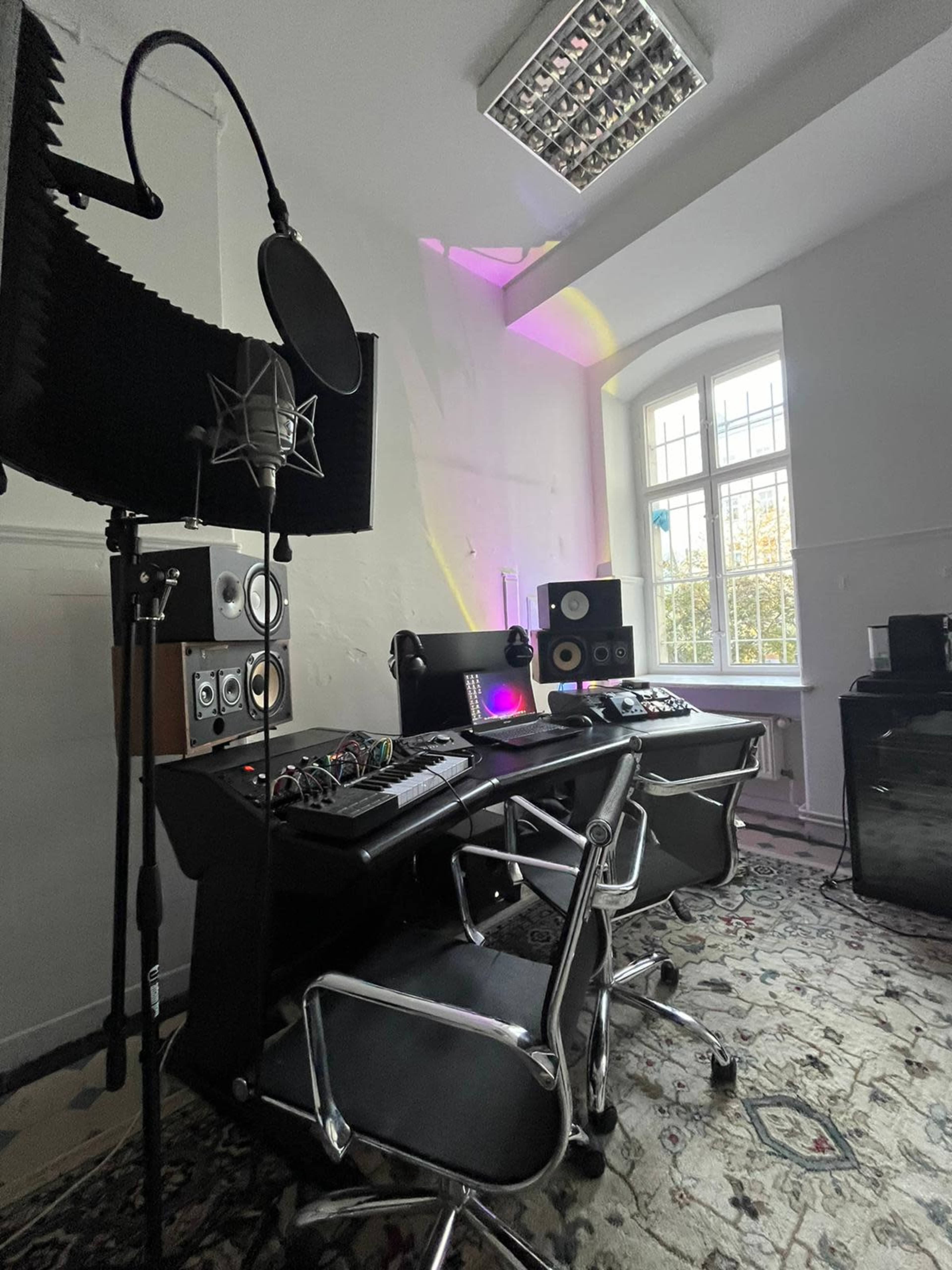 A modern music studio features a microphone, audio equipment, and two workstations set against a bright window.