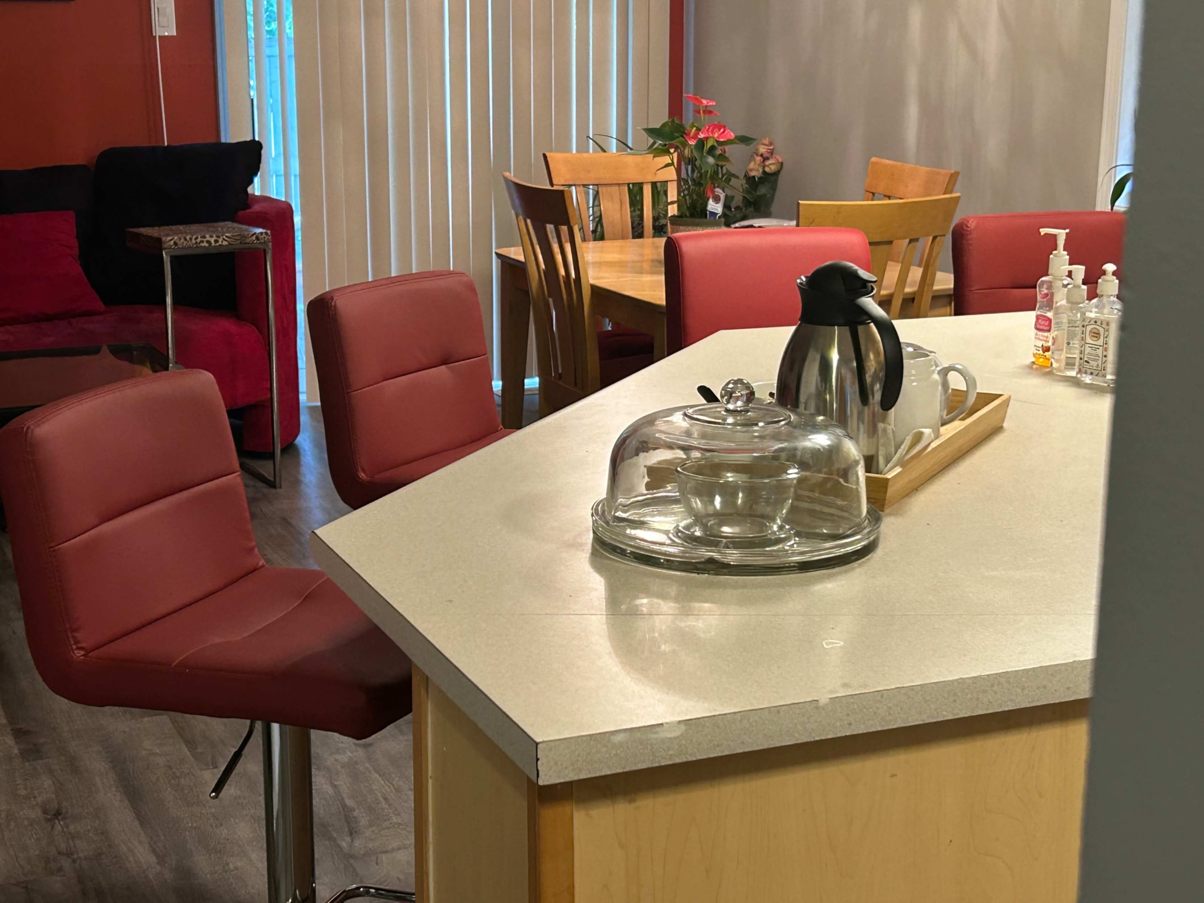The image shows a kitchen area featuring a countertop with a glass serving dome, a coffee pot, and several red chairs arranged around the space.