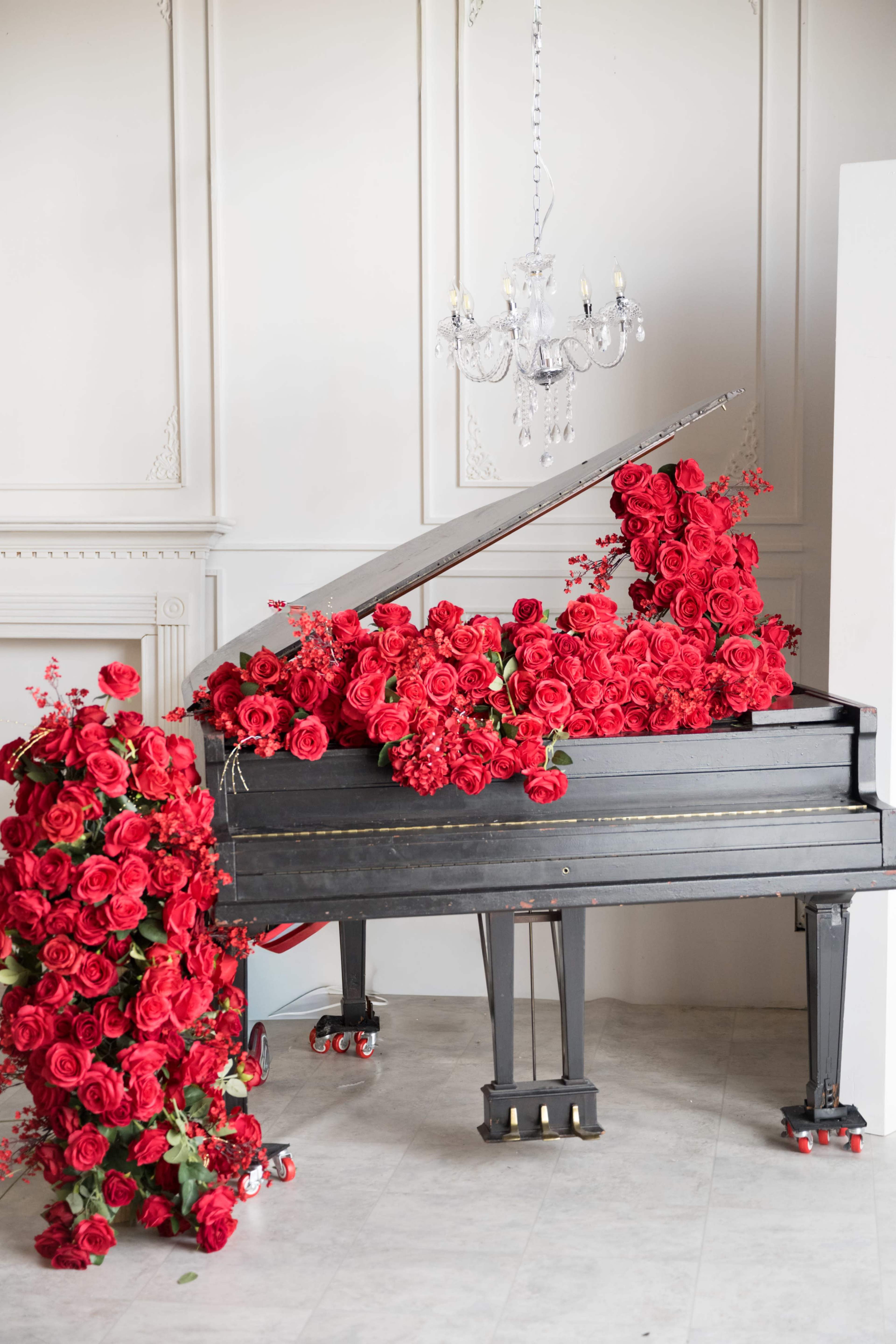 A grand piano is covered with an abundance of red roses, with a chandelier overhead in a well-lit room.