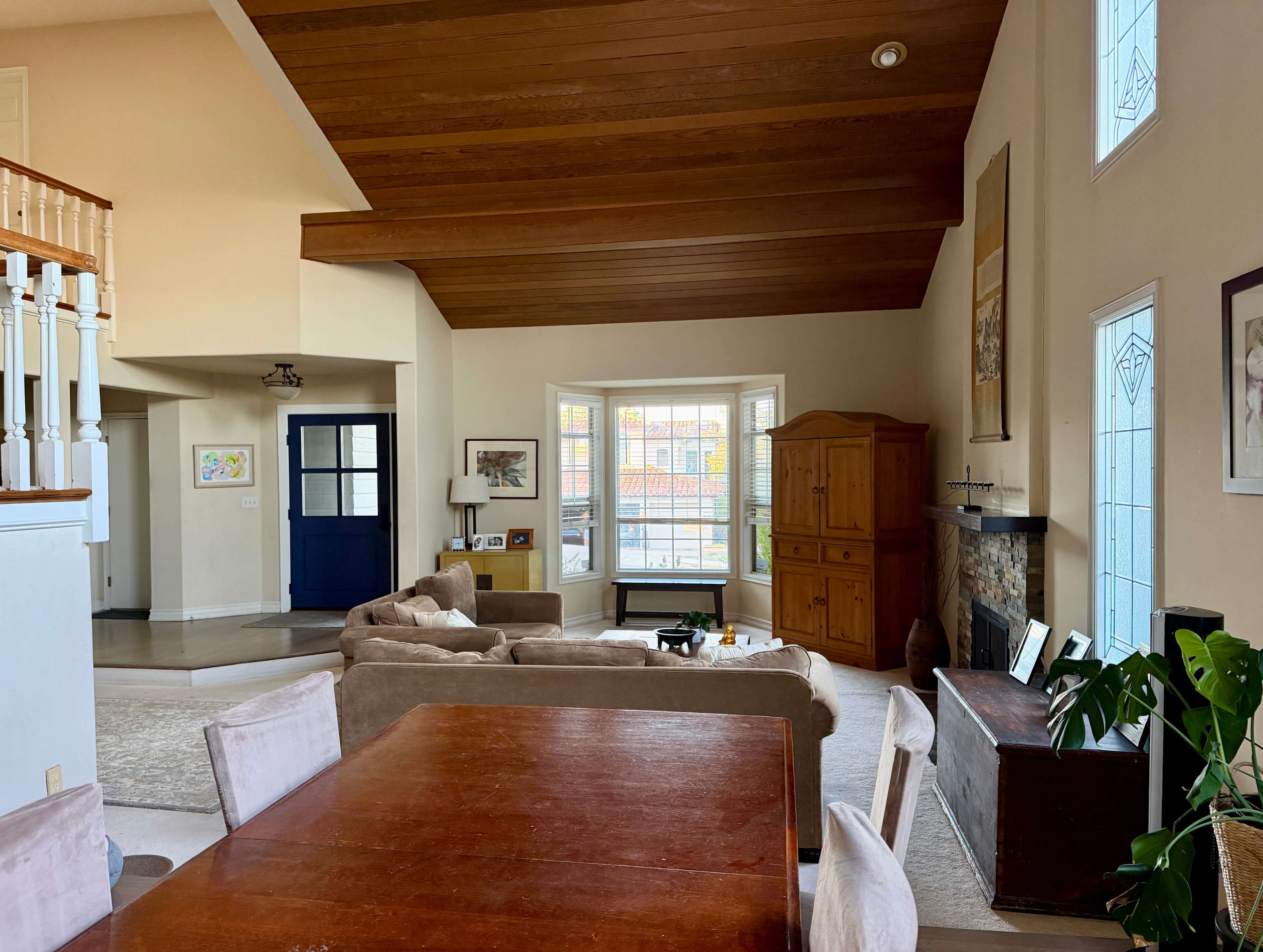 The image shows a spacious living area with a high wooden ceiling, a bay window, comfortable seating, and a wooden dining table.