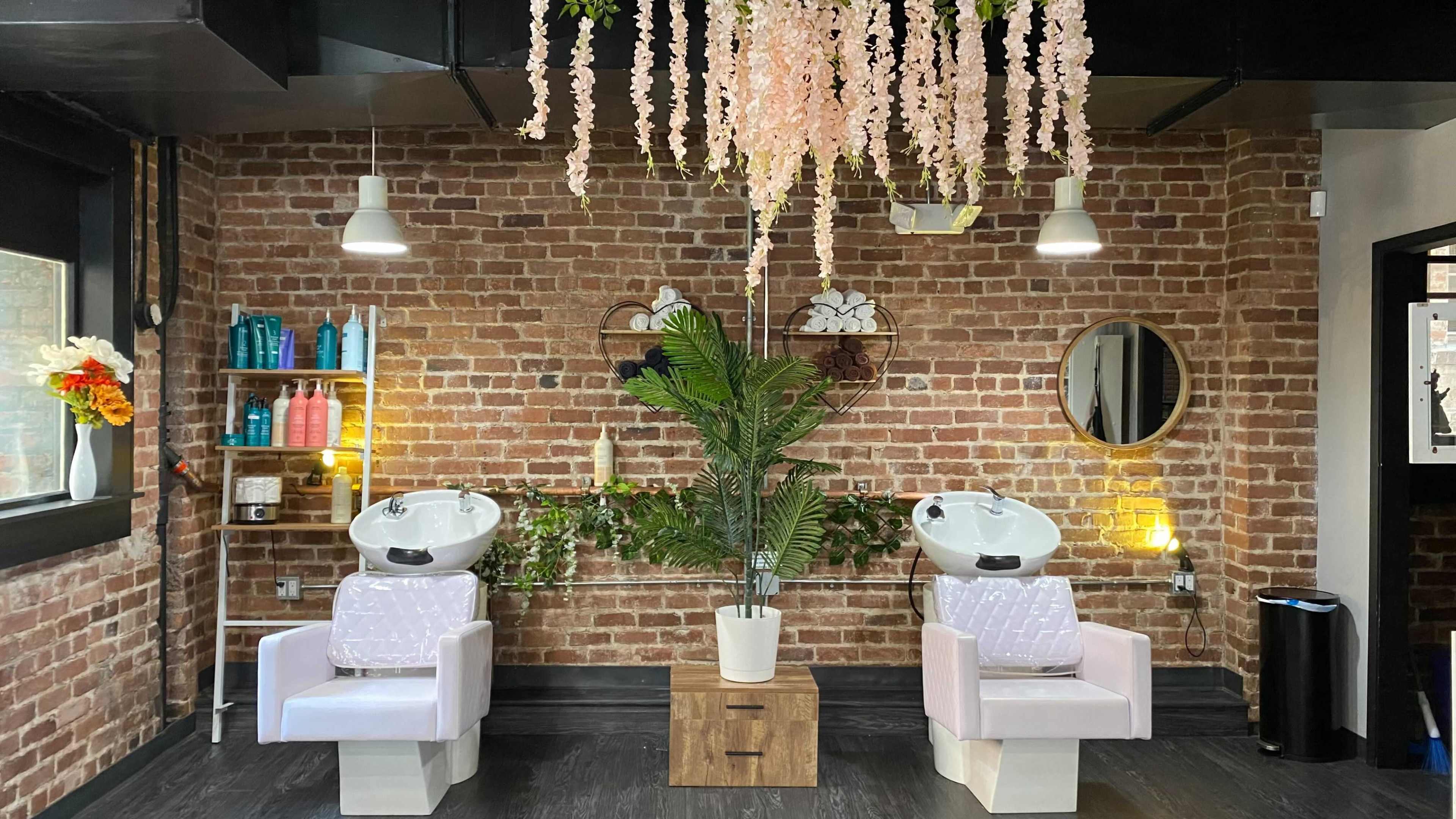 A salon interior featuring two hair washing stations with backlit mirrors, surrounded by a brick wall and adorned with hanging flowers and plants.