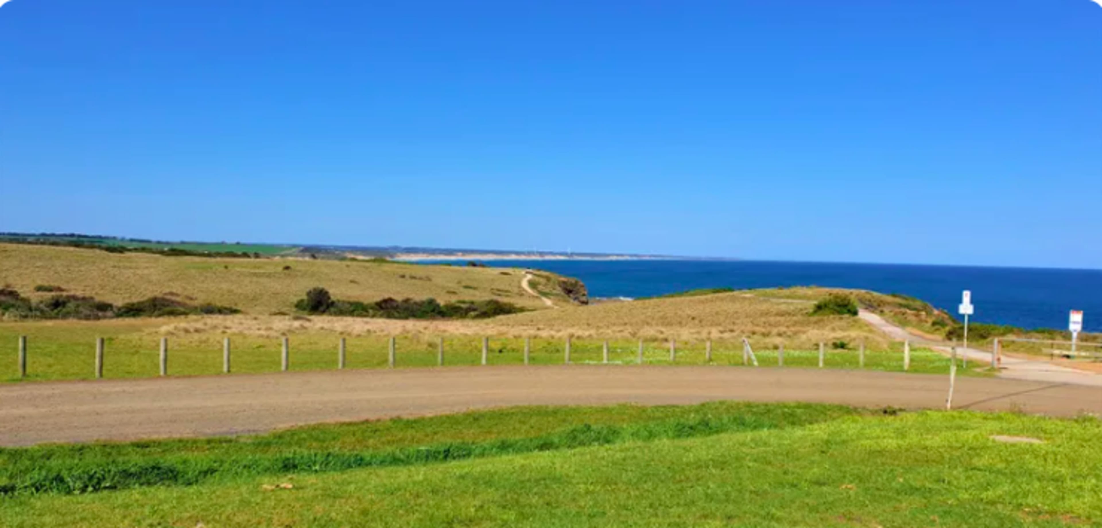 A wide view of a coastal landscape featuring grassy hills, a dirt road, and the ocean in the background.