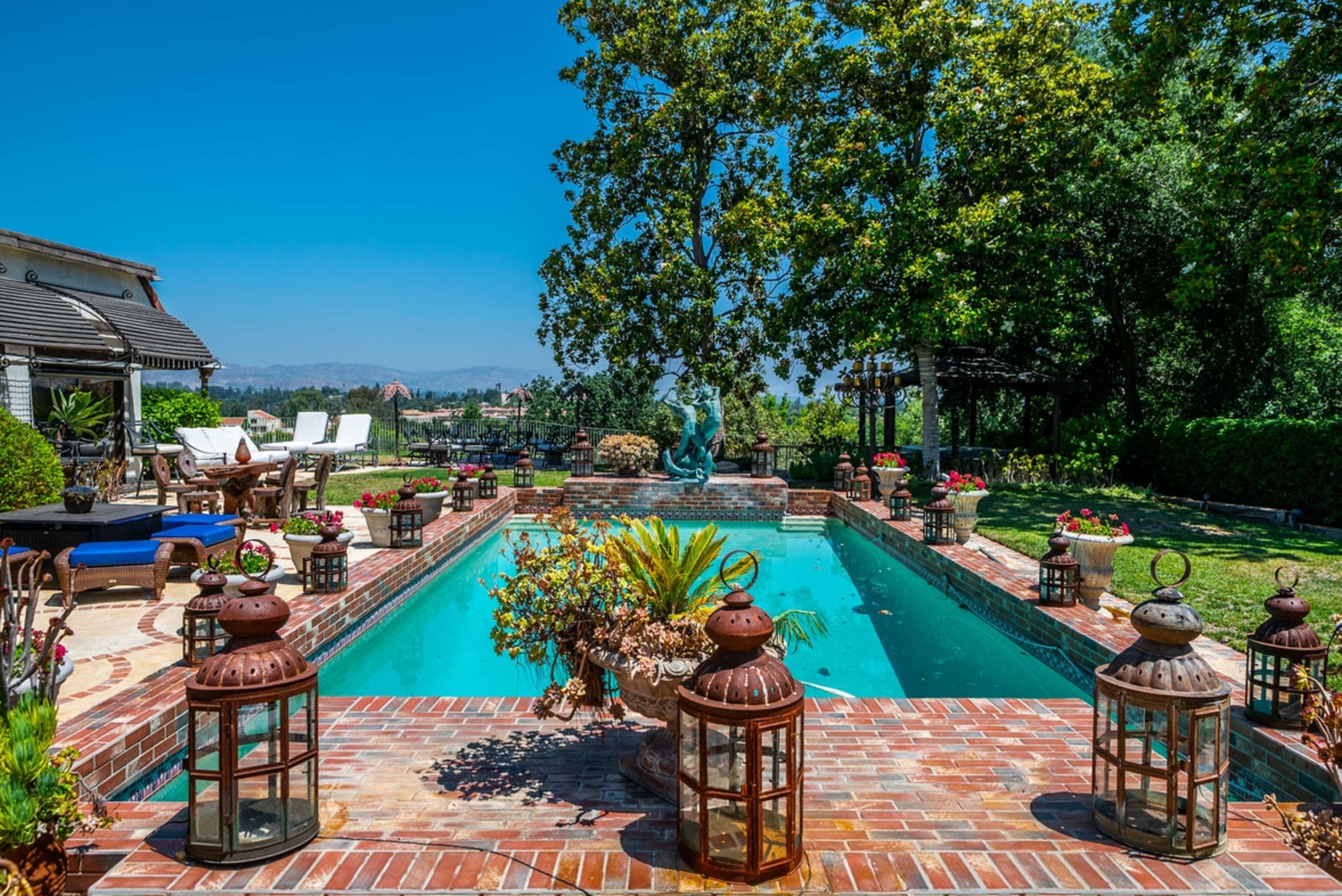 The image shows a backyard with a rectangular swimming pool surrounded by brick paving, lanterns, and lush greenery.