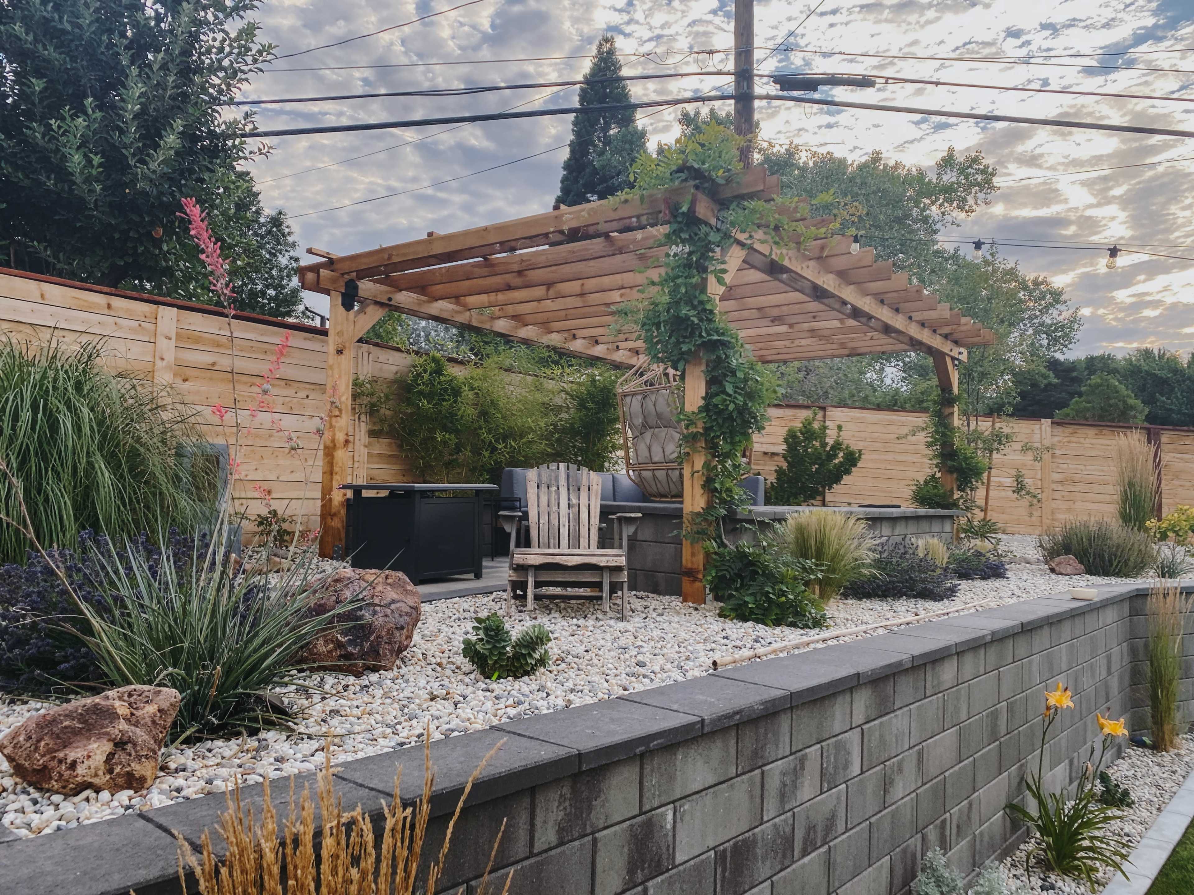 A wooden pergola with seating overlooks a landscaped garden featuring gravel, plants, and a BBQ area.