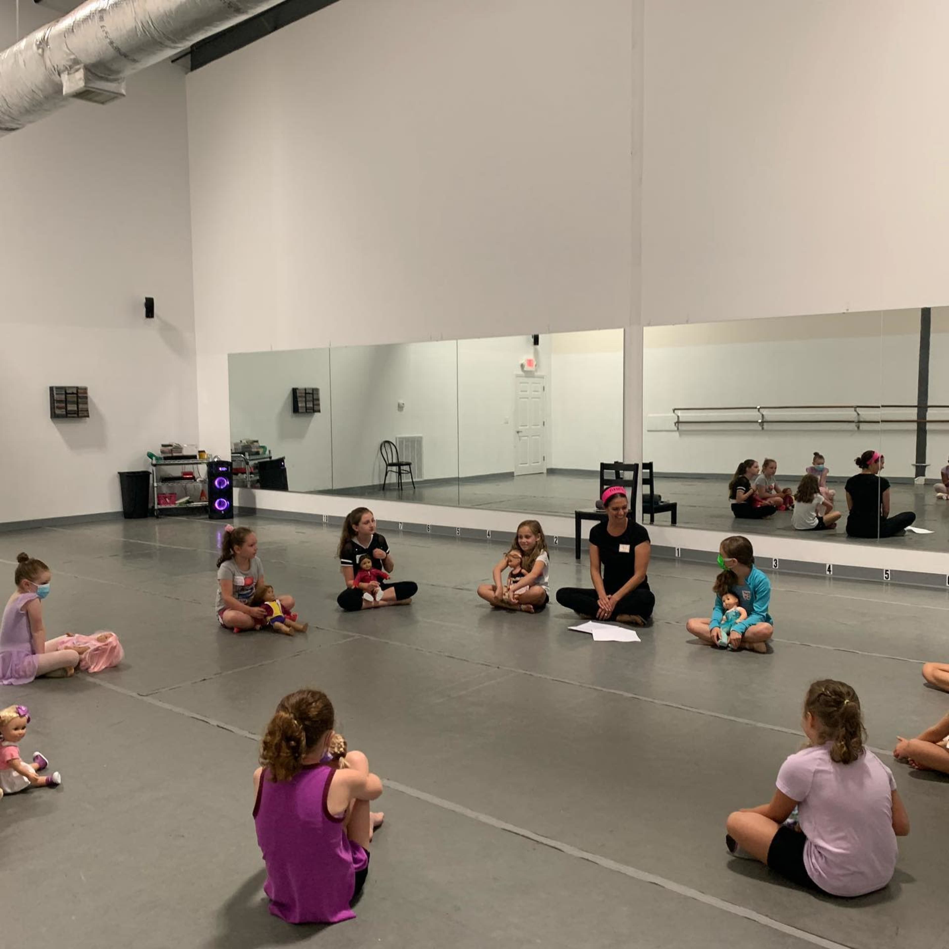 A group of children sits on the floor in a dance studio while an instructor leads a session in front of a mirror.