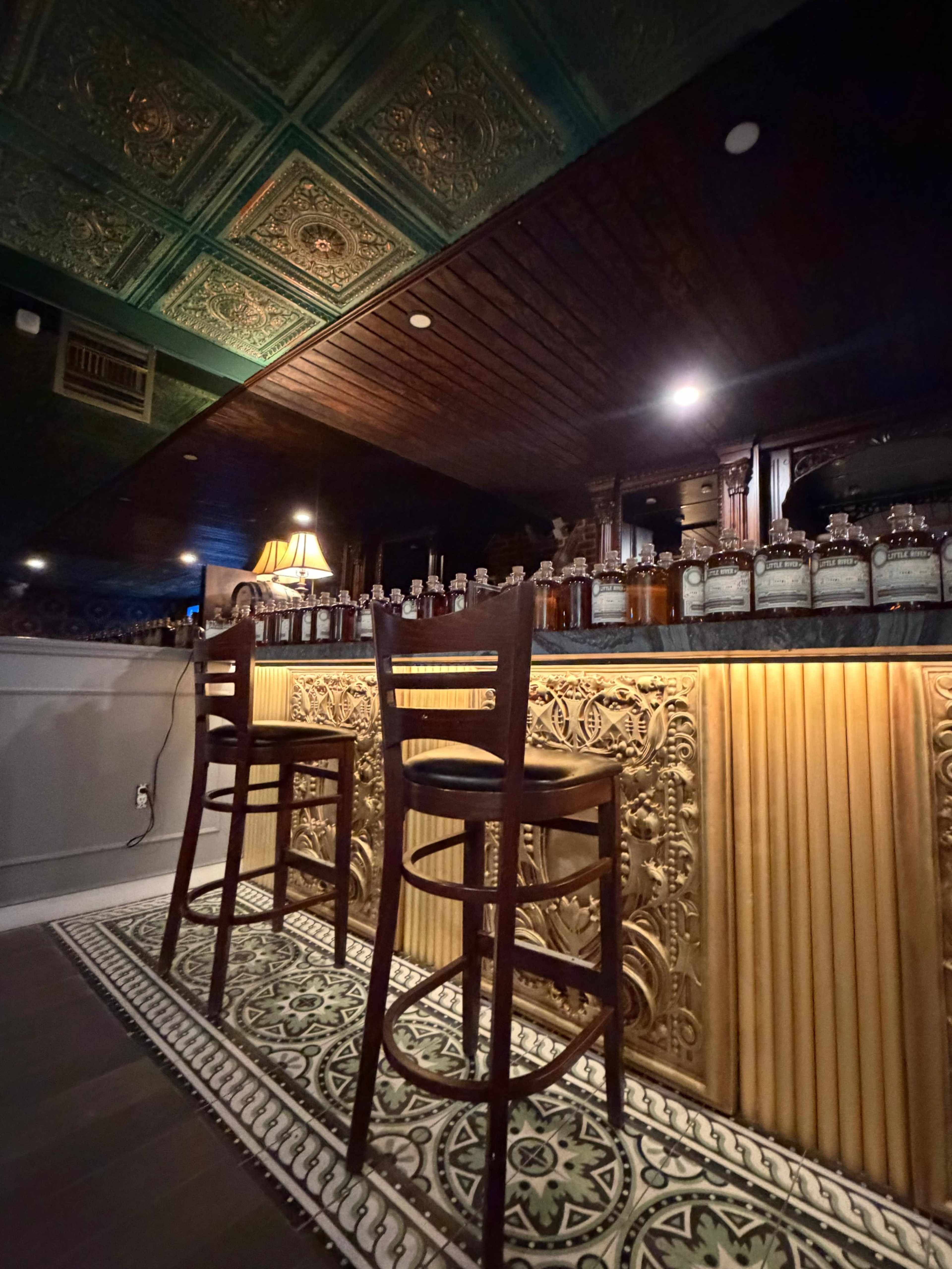 The image shows a bar area with two empty wooden stools and a shelf lined with bottles, all beneath an ornate ceiling design and warm lighting.