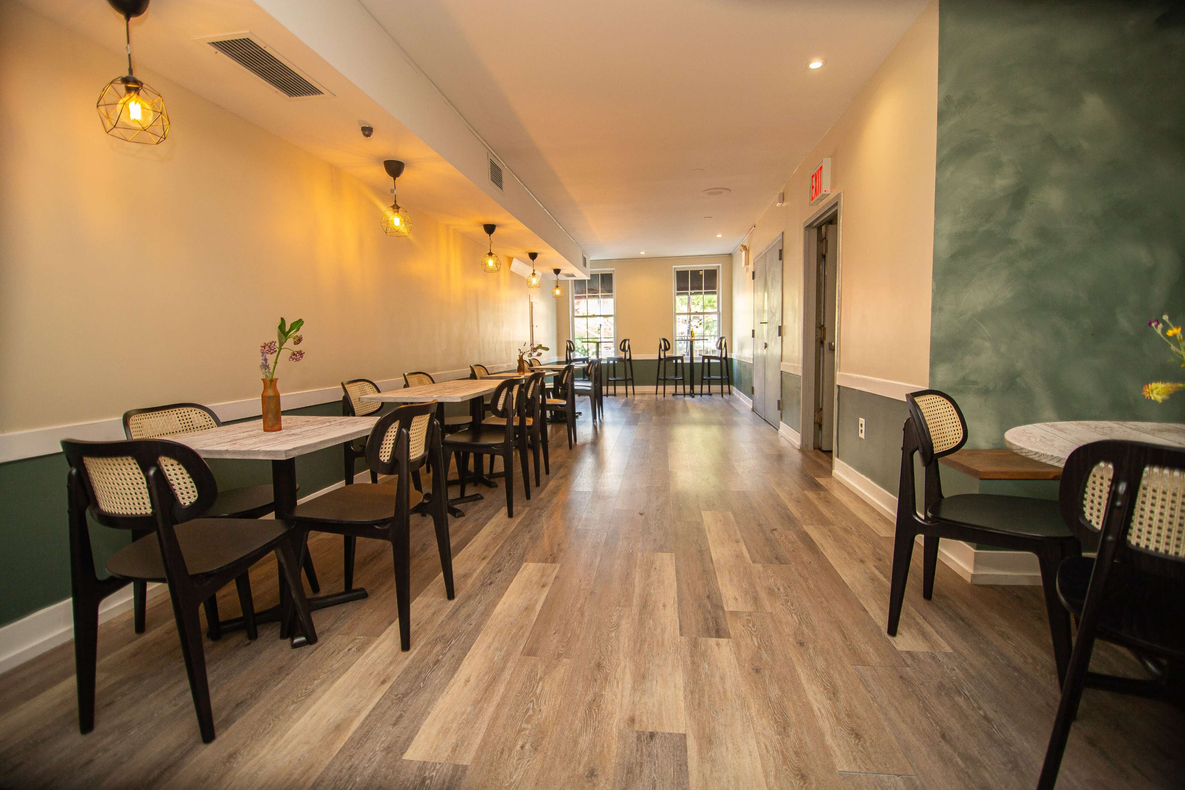 The image shows a modern, well-lit restaurant interior with wooden flooring, tables, and chairs arranged along a narrow aisle.