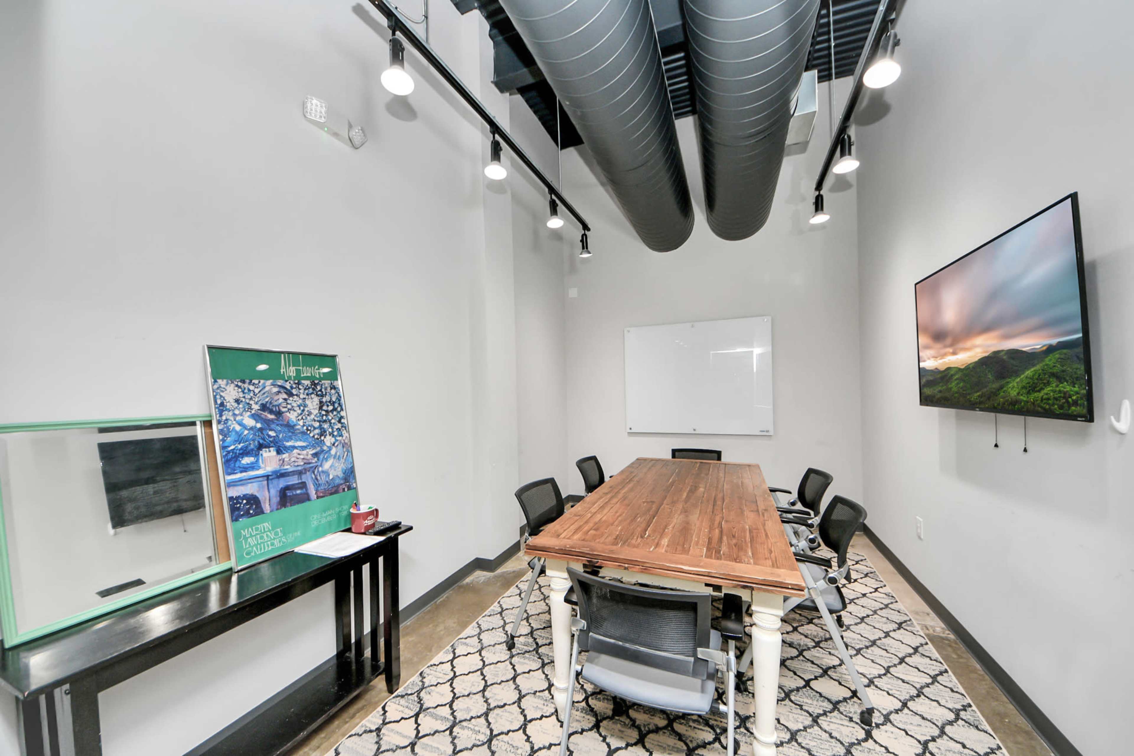 A modern conference room features a large wooden table, black chairs, and wall-mounted technology, with ductwork exposed on the ceiling.