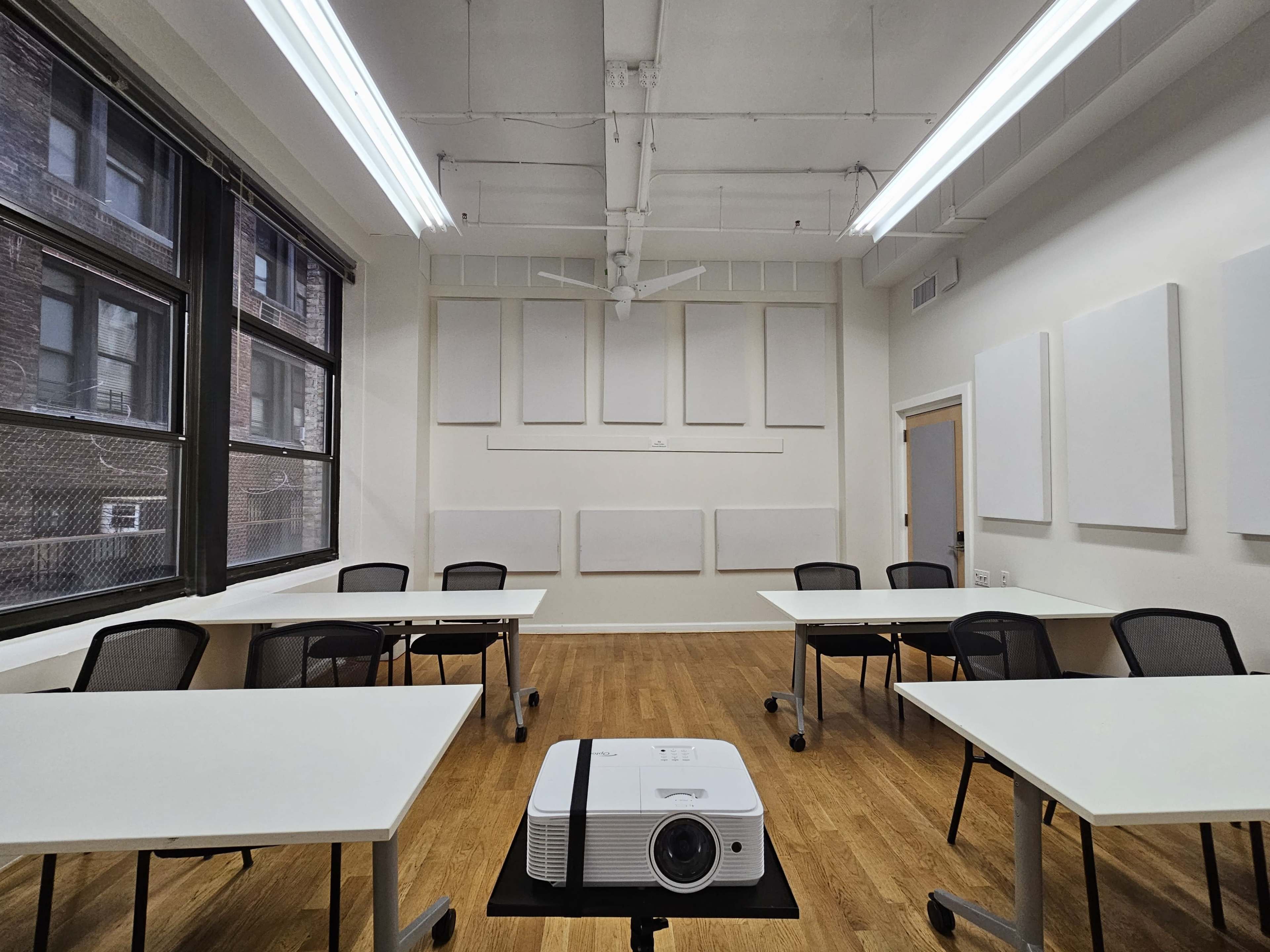 A classroom is arranged with several white tables and chairs, featuring a ceiling-mounted projector at the center.