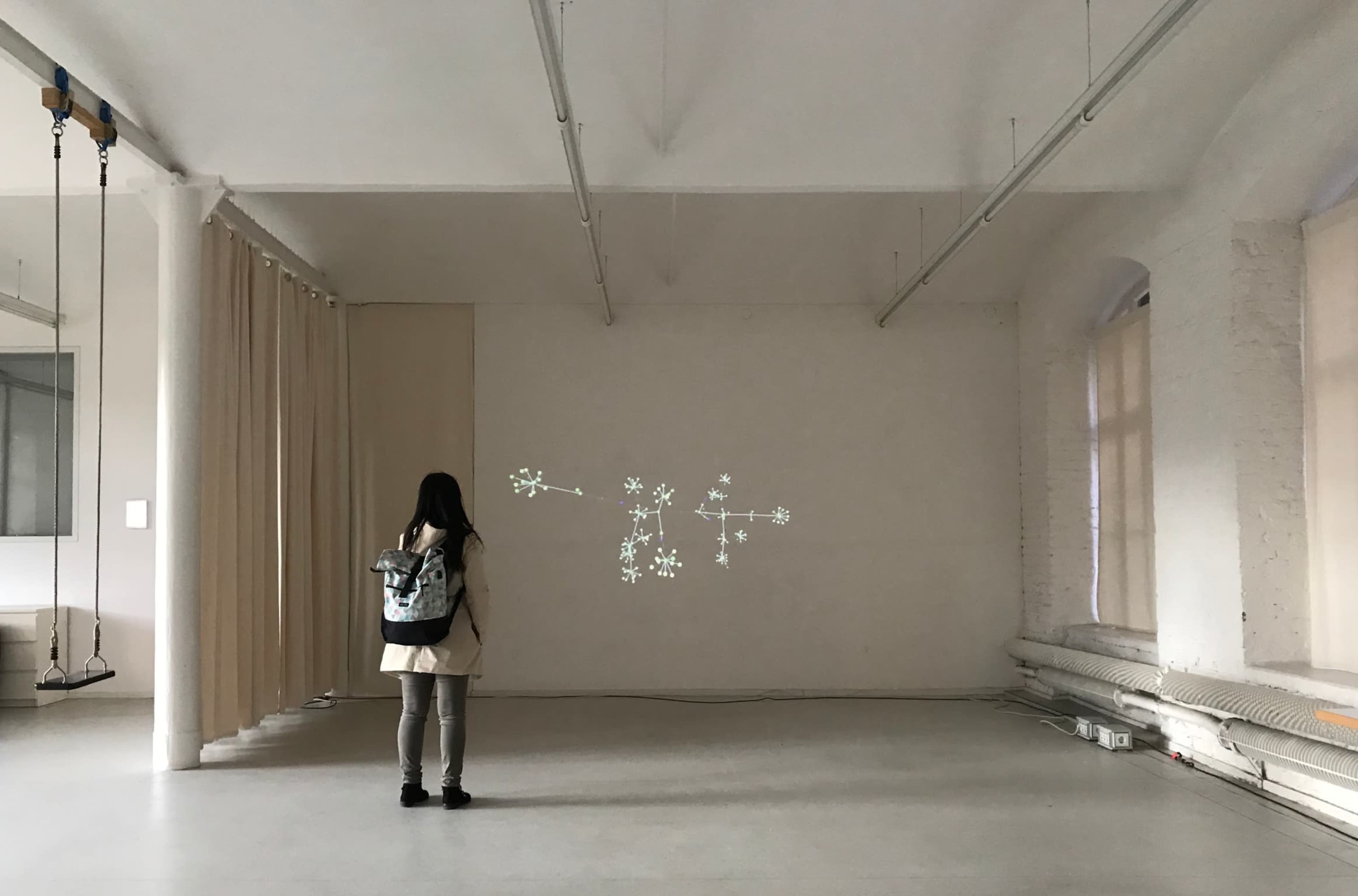 A person with a backpack stands in a spacious, minimally furnished room, looking at a projected display of stars on a blank wall.