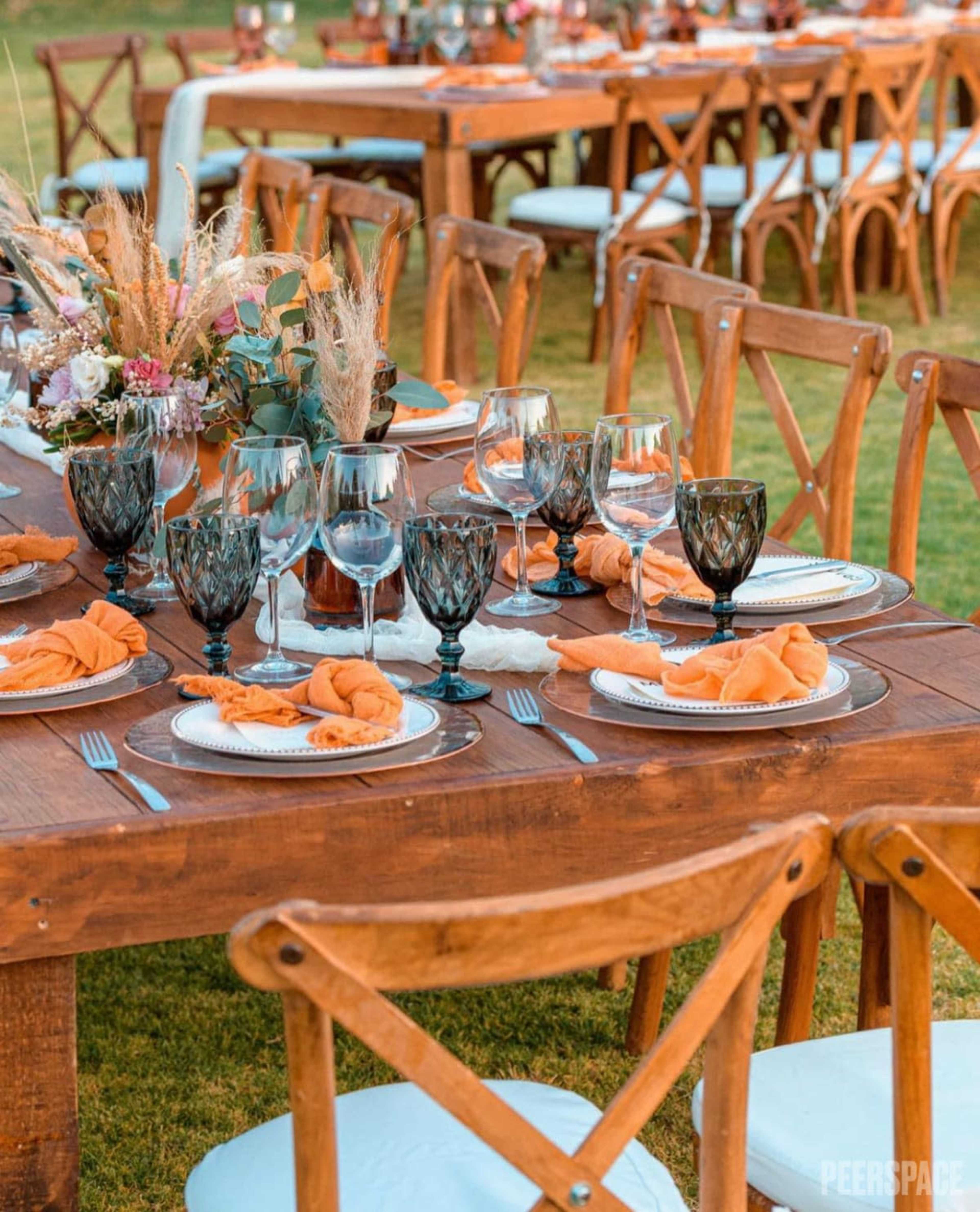 A long wooden dining table is set outdoors with elegant tableware, including glassware and neatly arranged napkins, surrounded by wooden chairs.