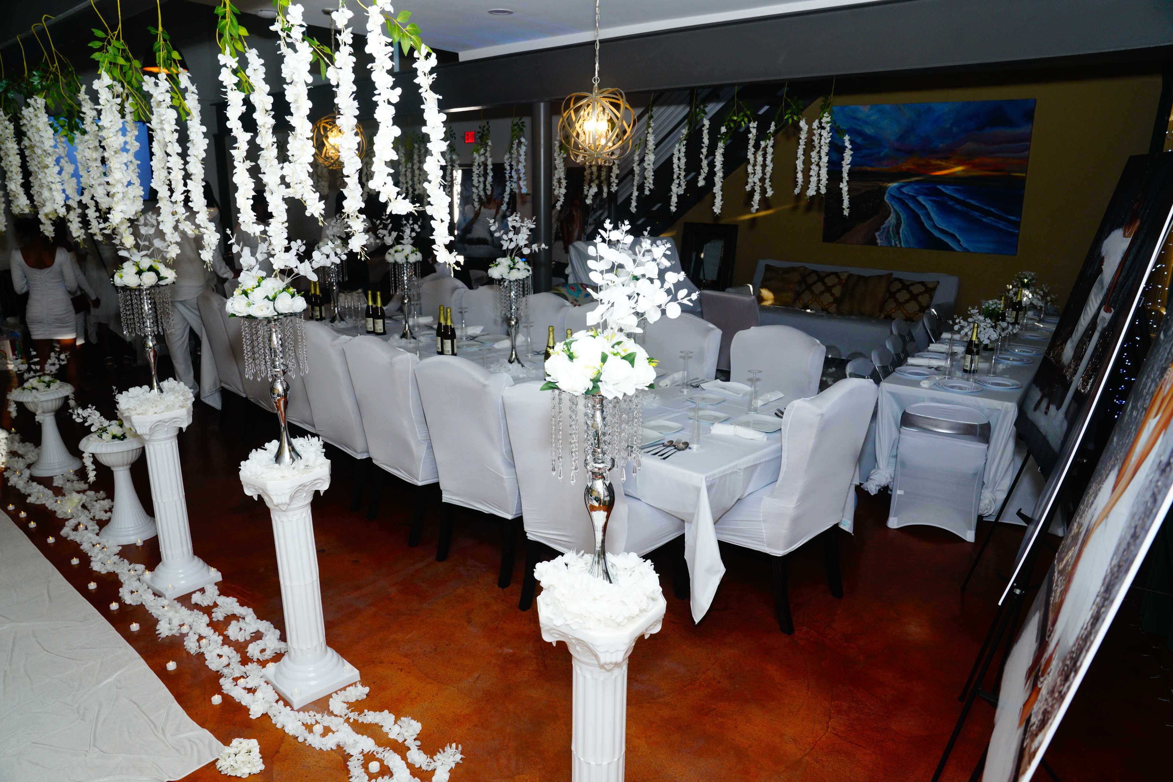 A elegantly decorated dining space with white-clothed tables set for an event, surrounded by floral arrangements and hanging decorations.