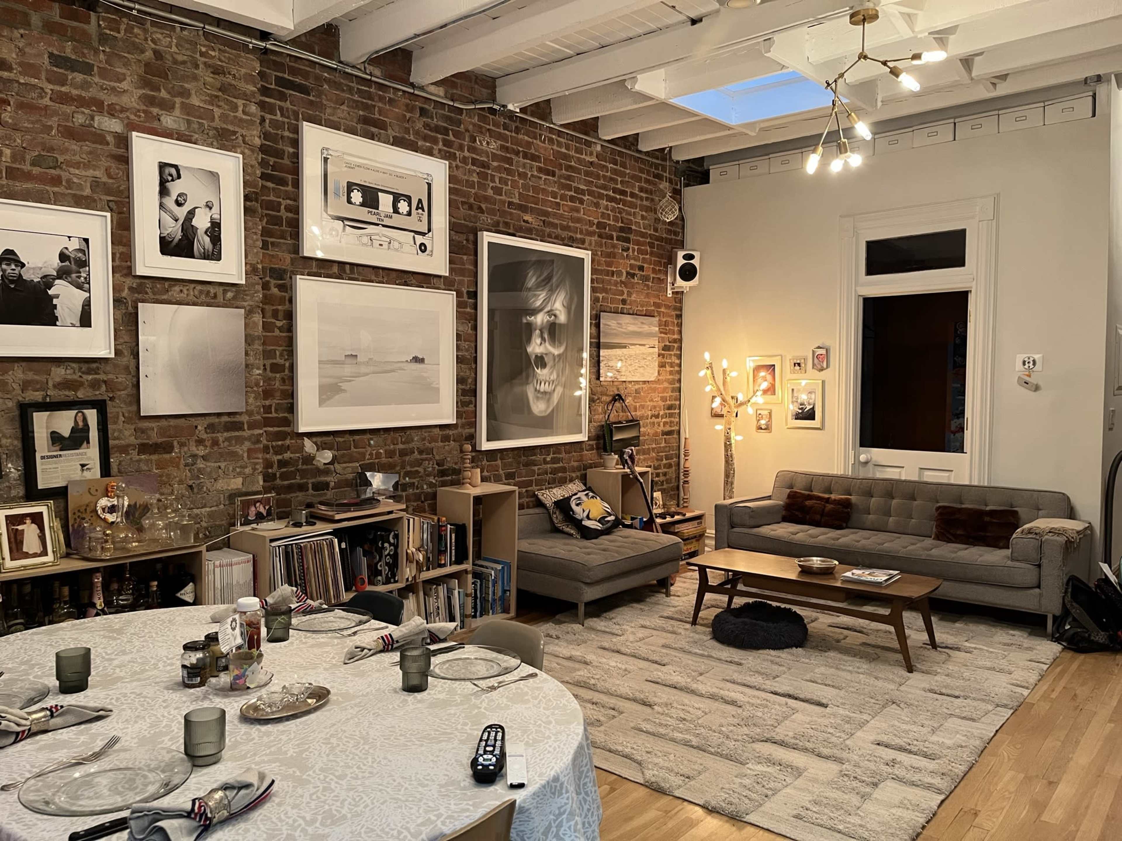 The image depicts a stylish living room and dining area with exposed brick walls, a large round dining table set for a meal, a cozy sofa, various artworks on the walls, and ambient lighting.