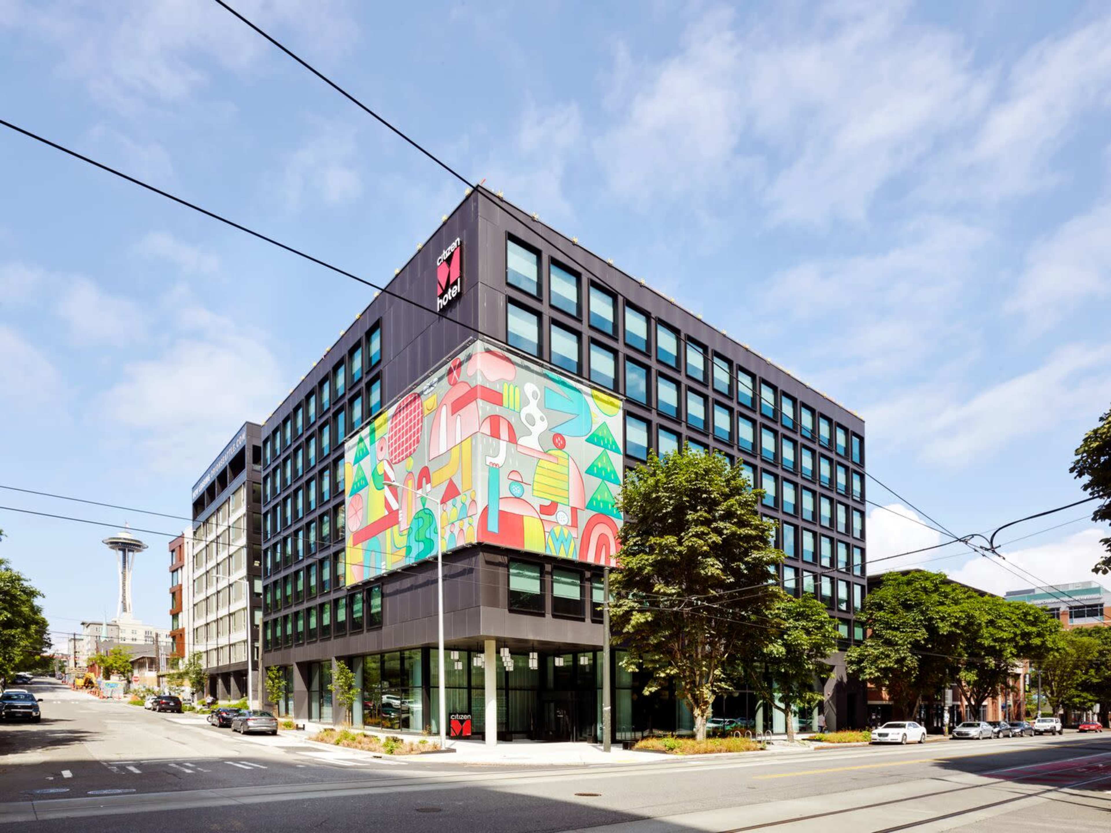 A modern, multi-story building features a large, colorful mural on its side, situated at an urban intersection with trees and the Space Needle visible in the background.