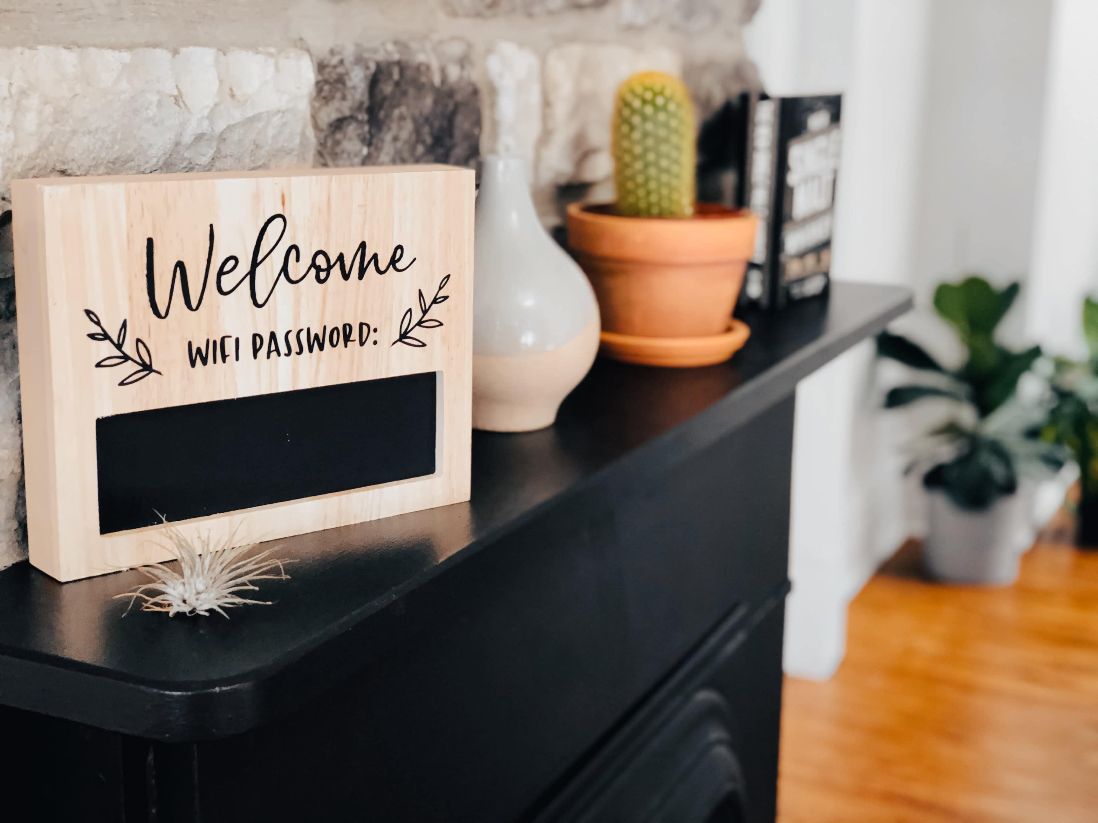 A wooden sign with "Welcome" and "WIFI Password:" is placed on a black mantel alongside a vase, a potted cactus, and a small air plant.