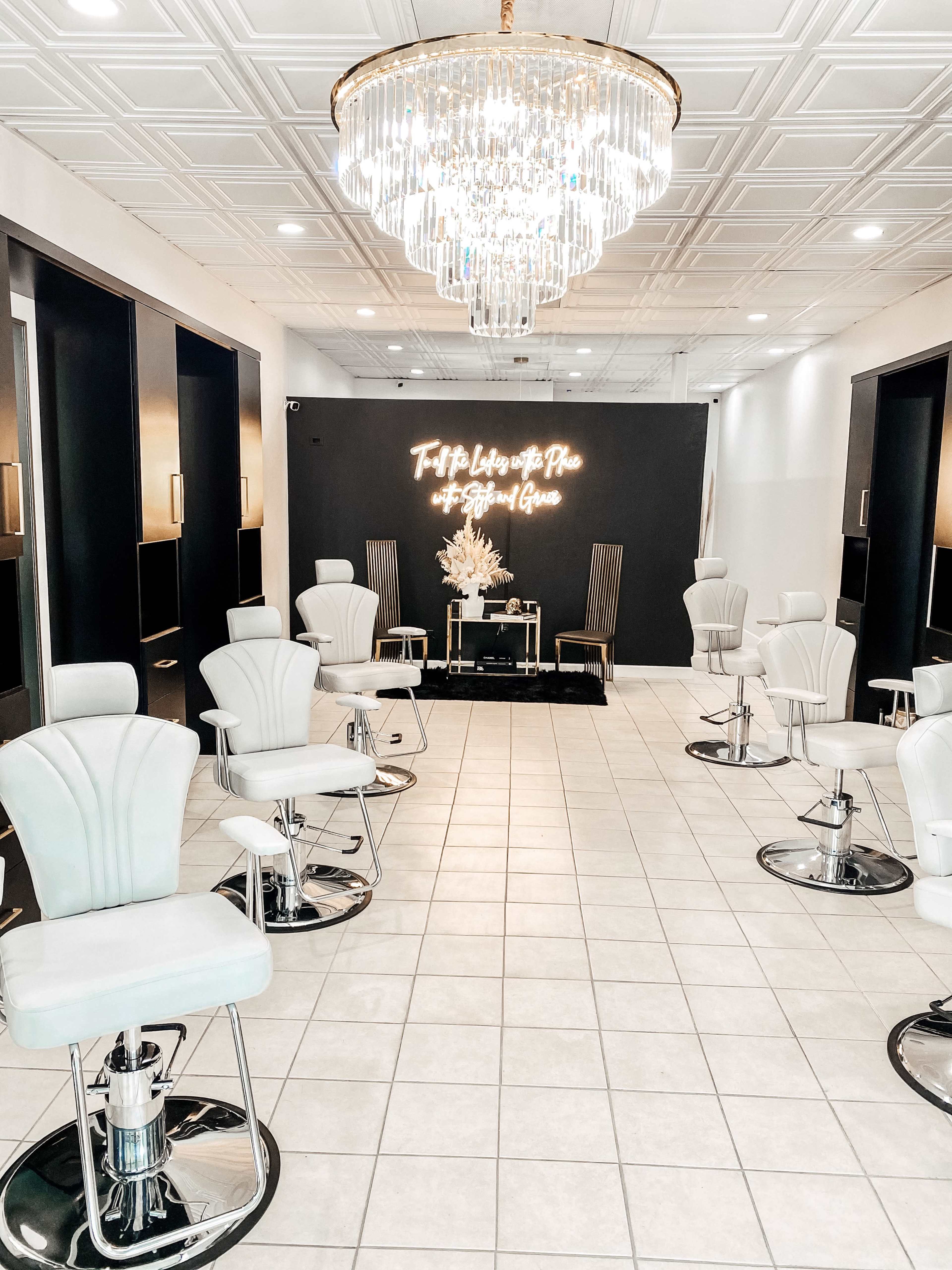 A modern salon interior featuring several white styling chairs arranged in front of a black wall with neon lighting and a chandelier above.