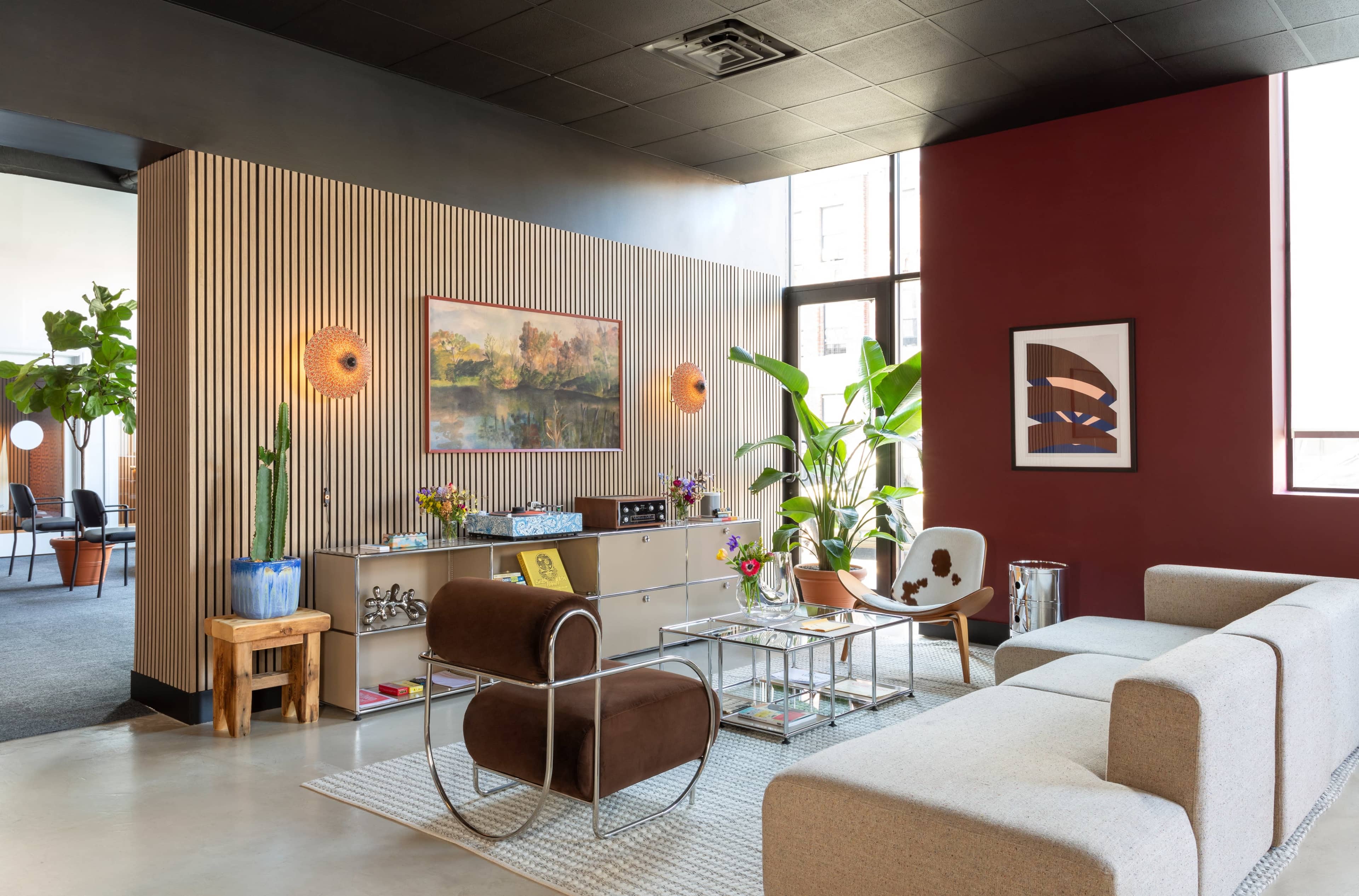 The image shows a modern living space with a light-colored sofa, a brown chair, a decorative sideboard, and various plants, framed artwork, and colorful decor.