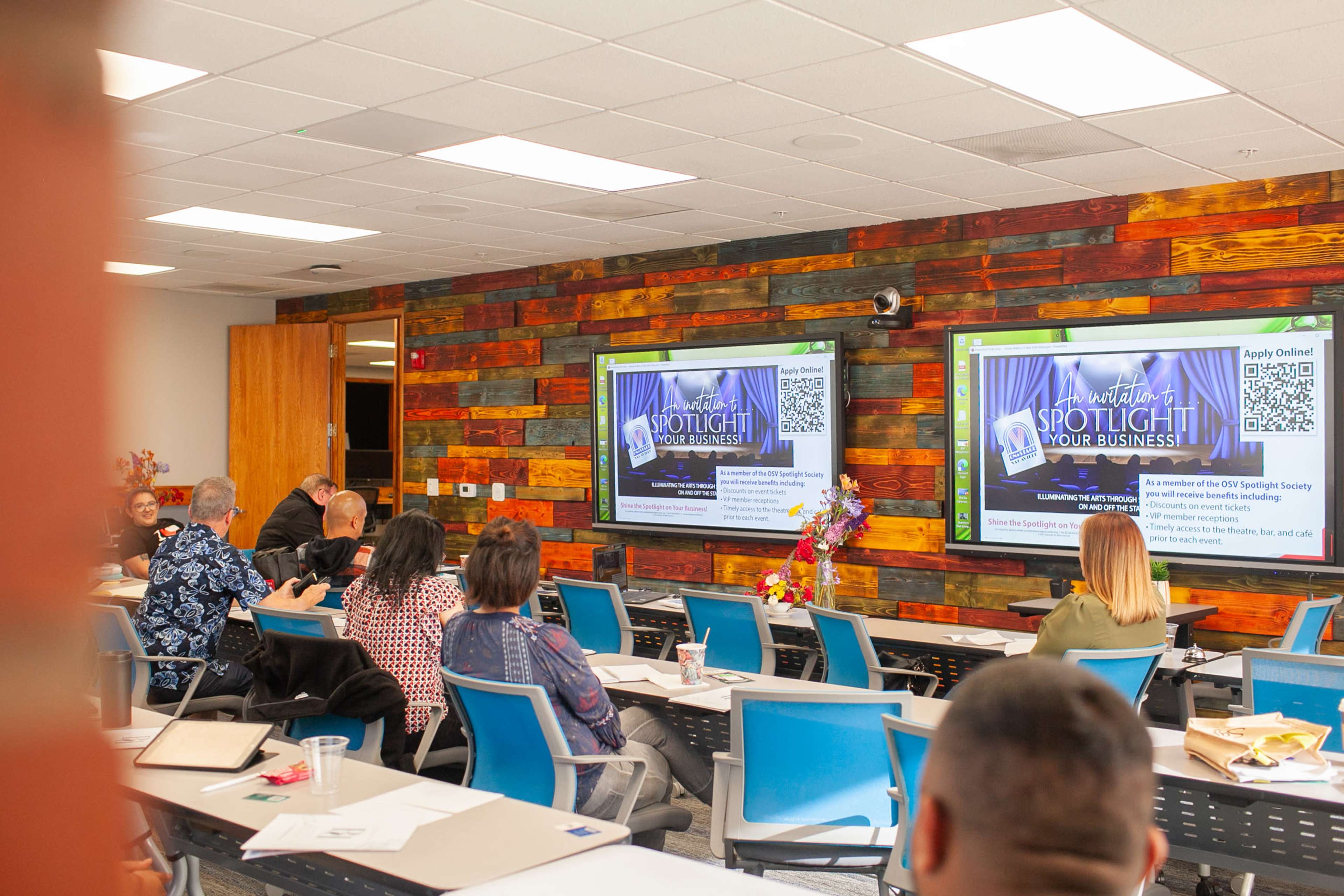A group of people are seated in a conference room with two large screens displaying a presentation about promoting local businesses.