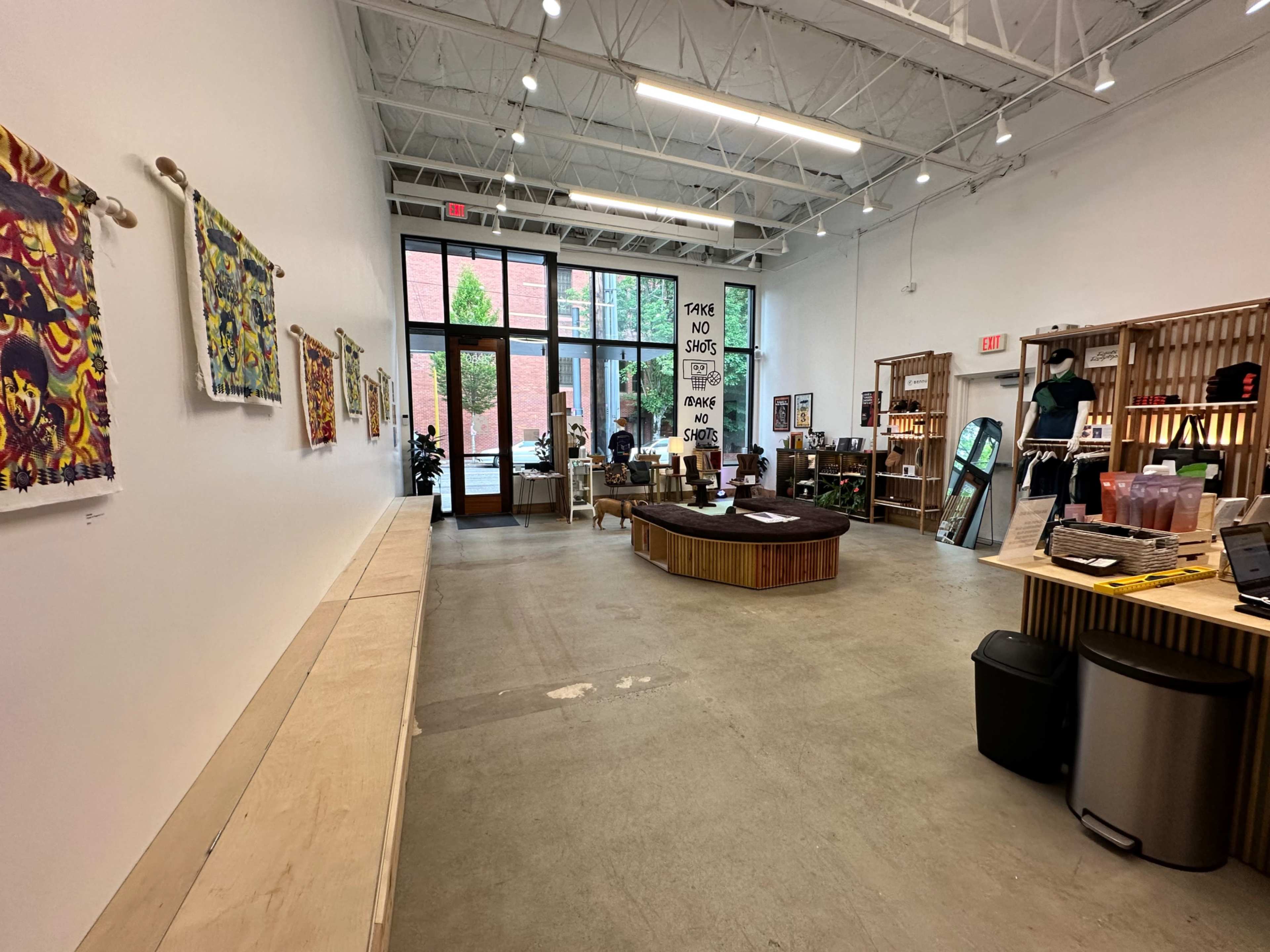 A spacious retail interior with wooden display furniture, colorful artwork on the walls, and a large entrance allowing natural light.