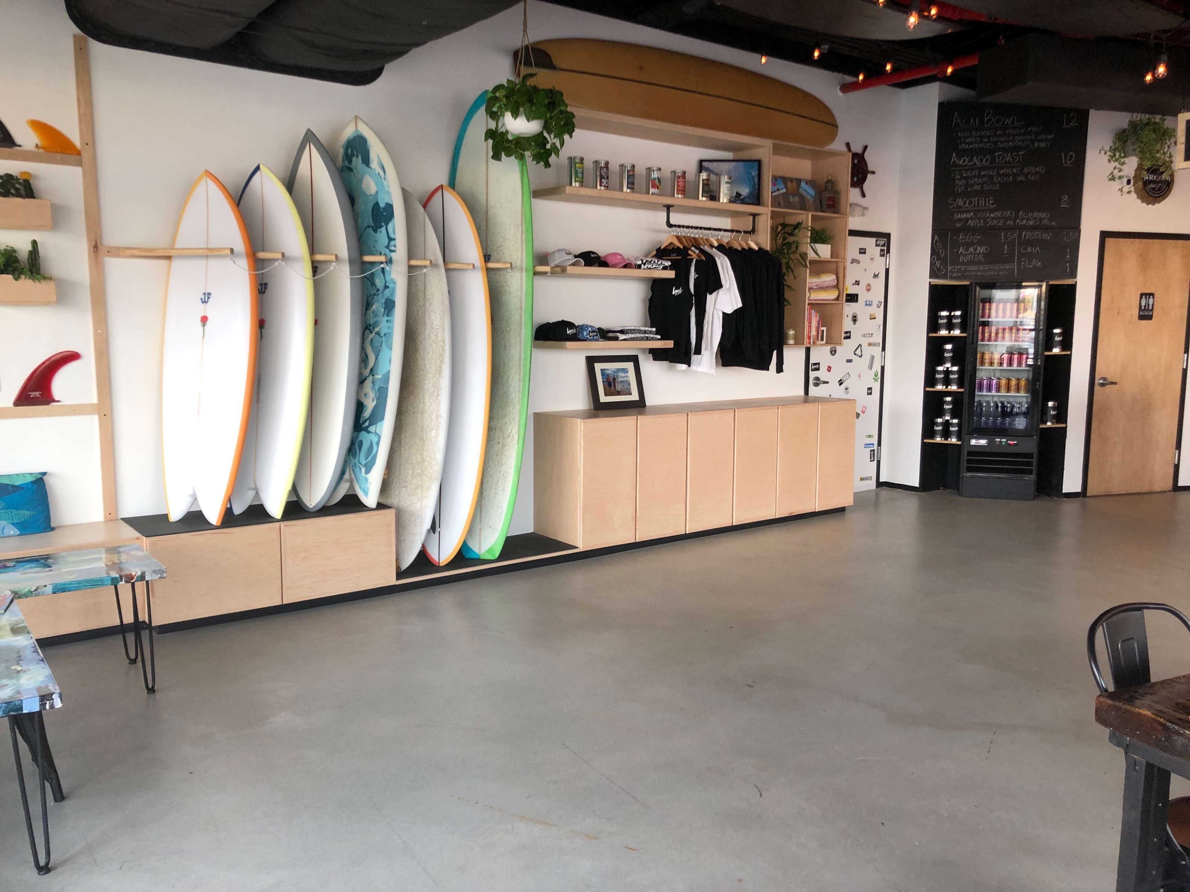 The image shows a surf shop interior with multiple surfboards displayed on a wall, clothing hanging on a rack, and a refrigerator against the back wall.