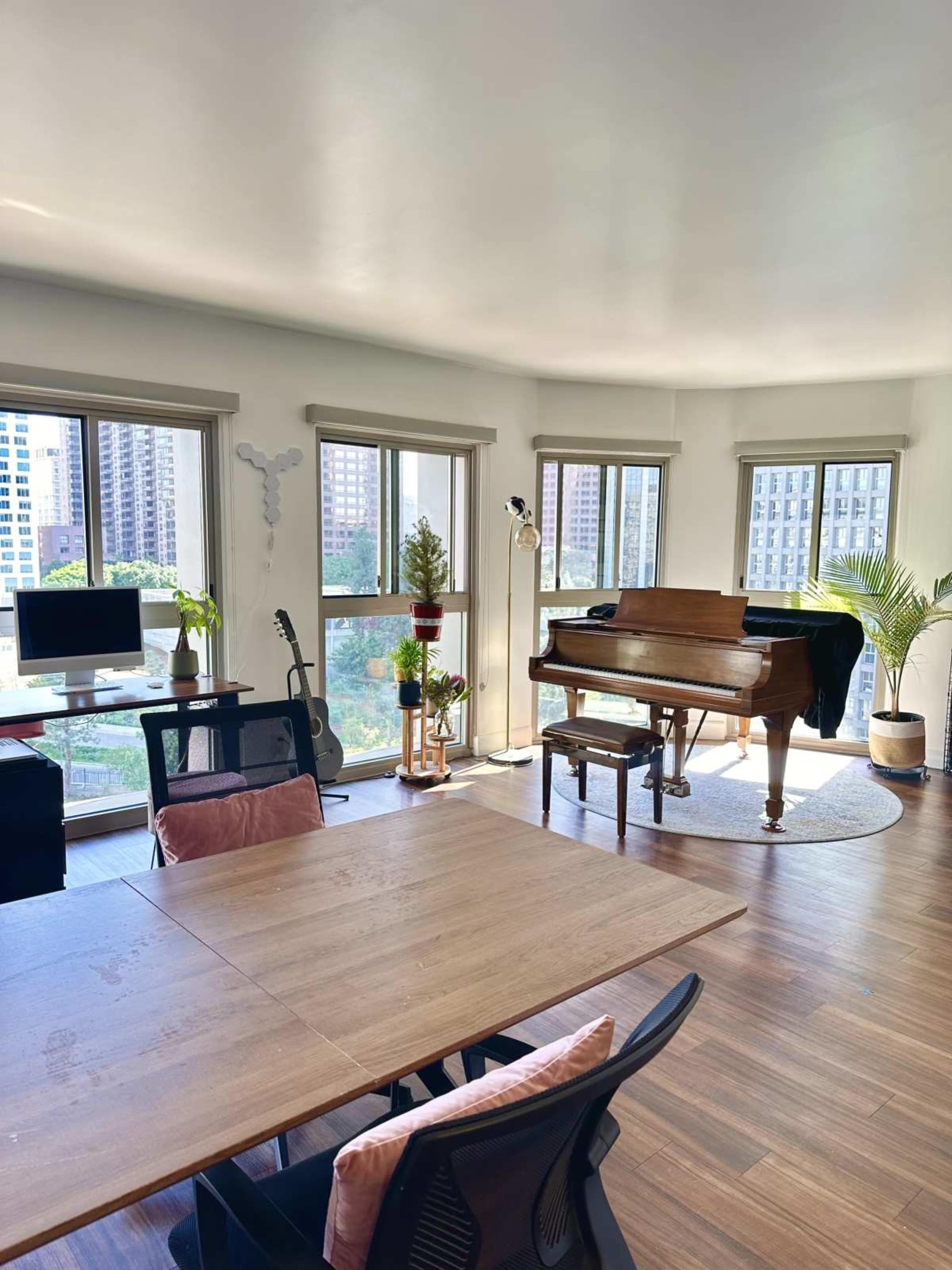 The image shows a sunlit modern living room featuring a grand piano, a dining table with chairs, a desk with a computer, and several plants near large windows.