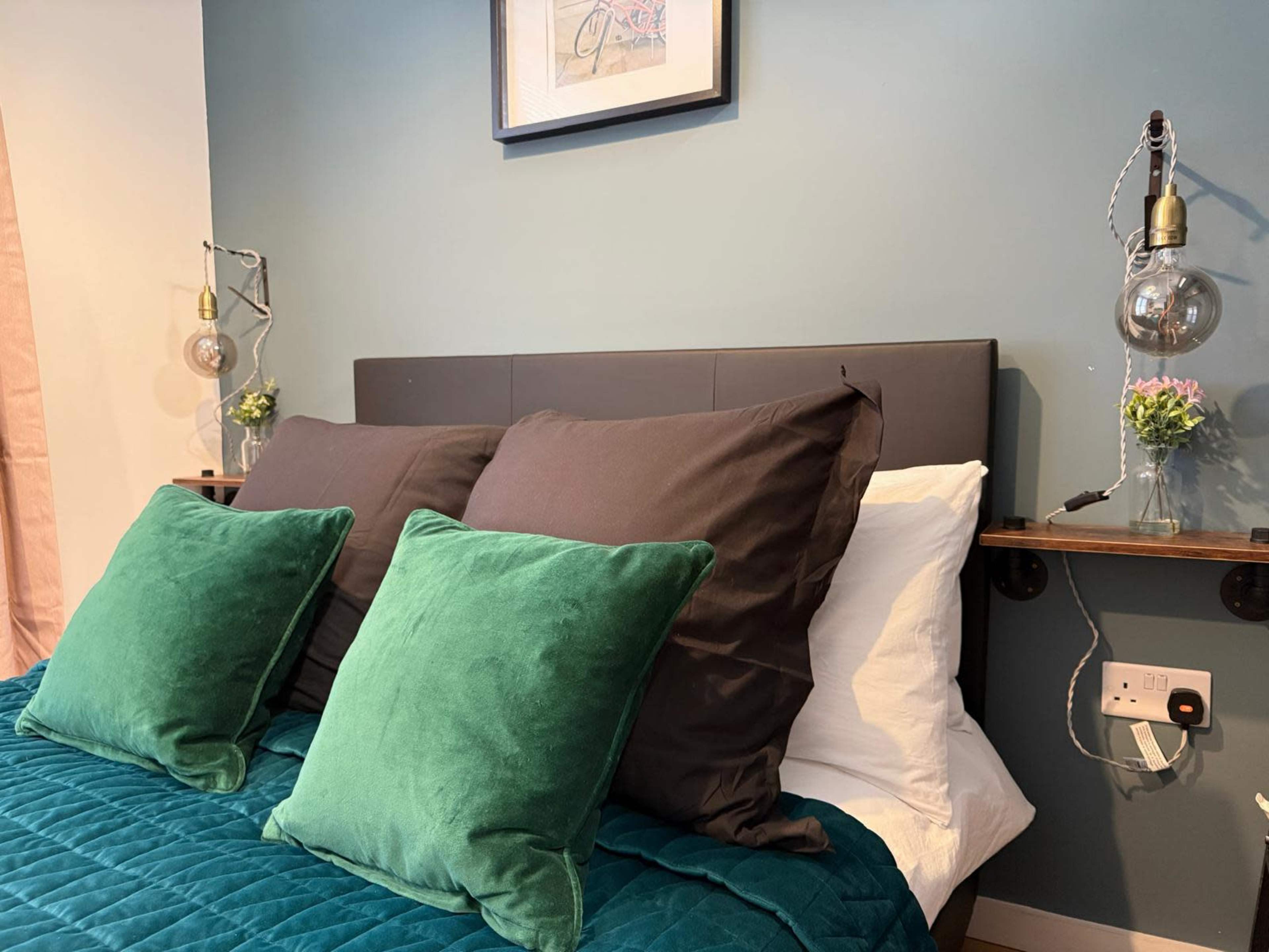 The image shows a neatly made bed with two dark pillows and two green velvet throw pillows, flanked by wall-mounted lamps and a small shelf with decorative items.