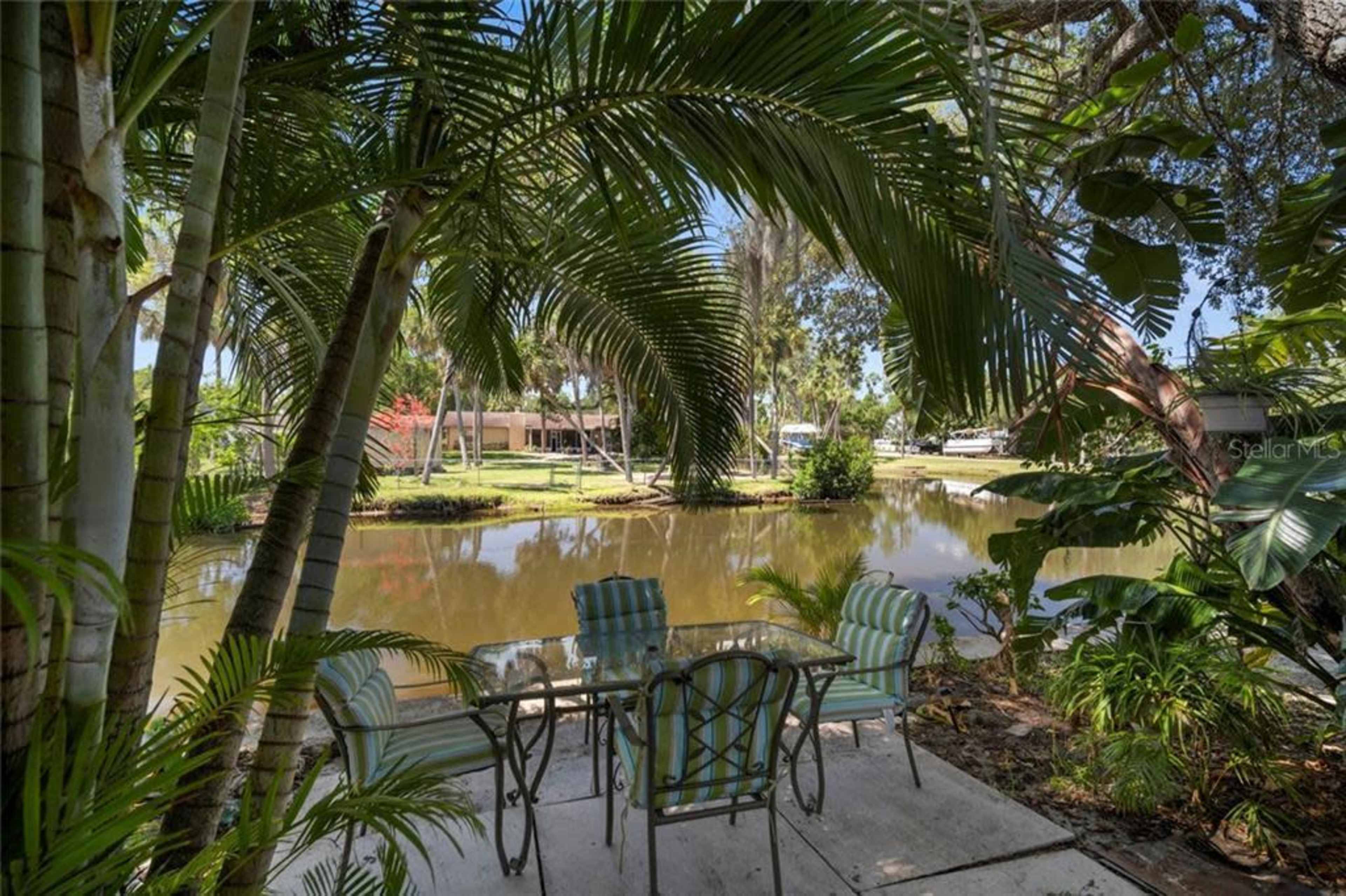 A table with chairs is set beside a calm body of water, surrounded by lush greenery and palm trees.