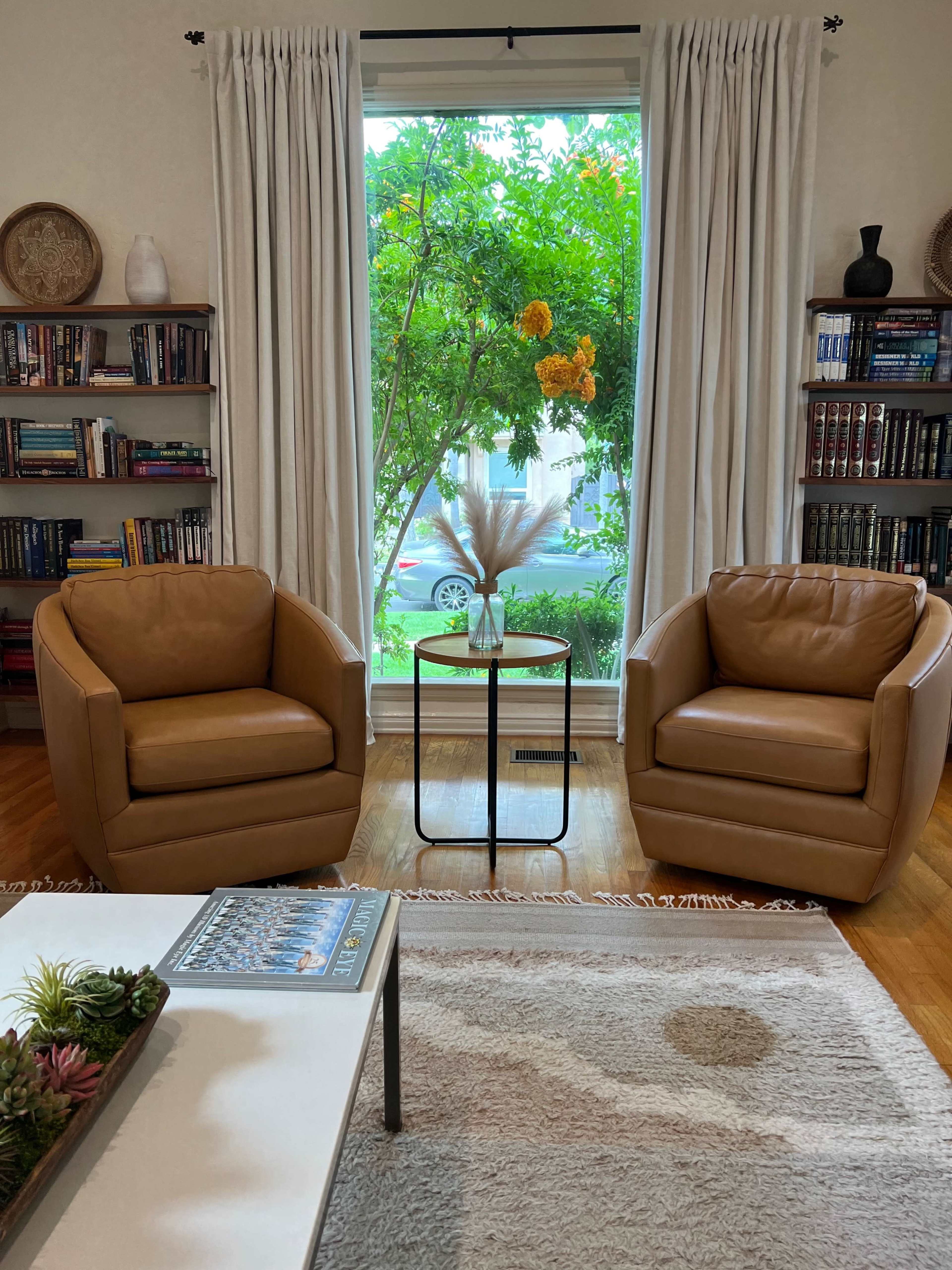 A cozy living room with two tan armchairs facing a small black table, surrounded by bookshelves and a large window that overlooks greenery.