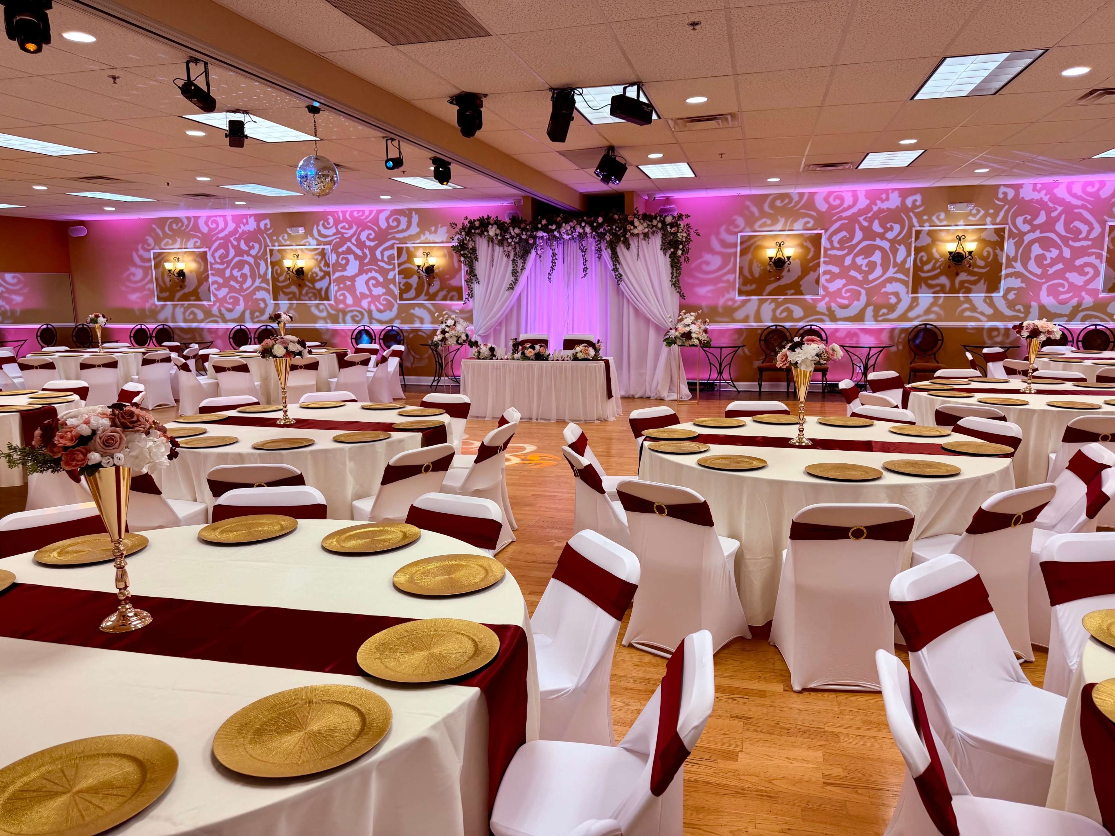 The image shows a decorated banquet hall with round tables covered in white tablecloths and burgundy runners, set with gold chargers and floral centerpieces.