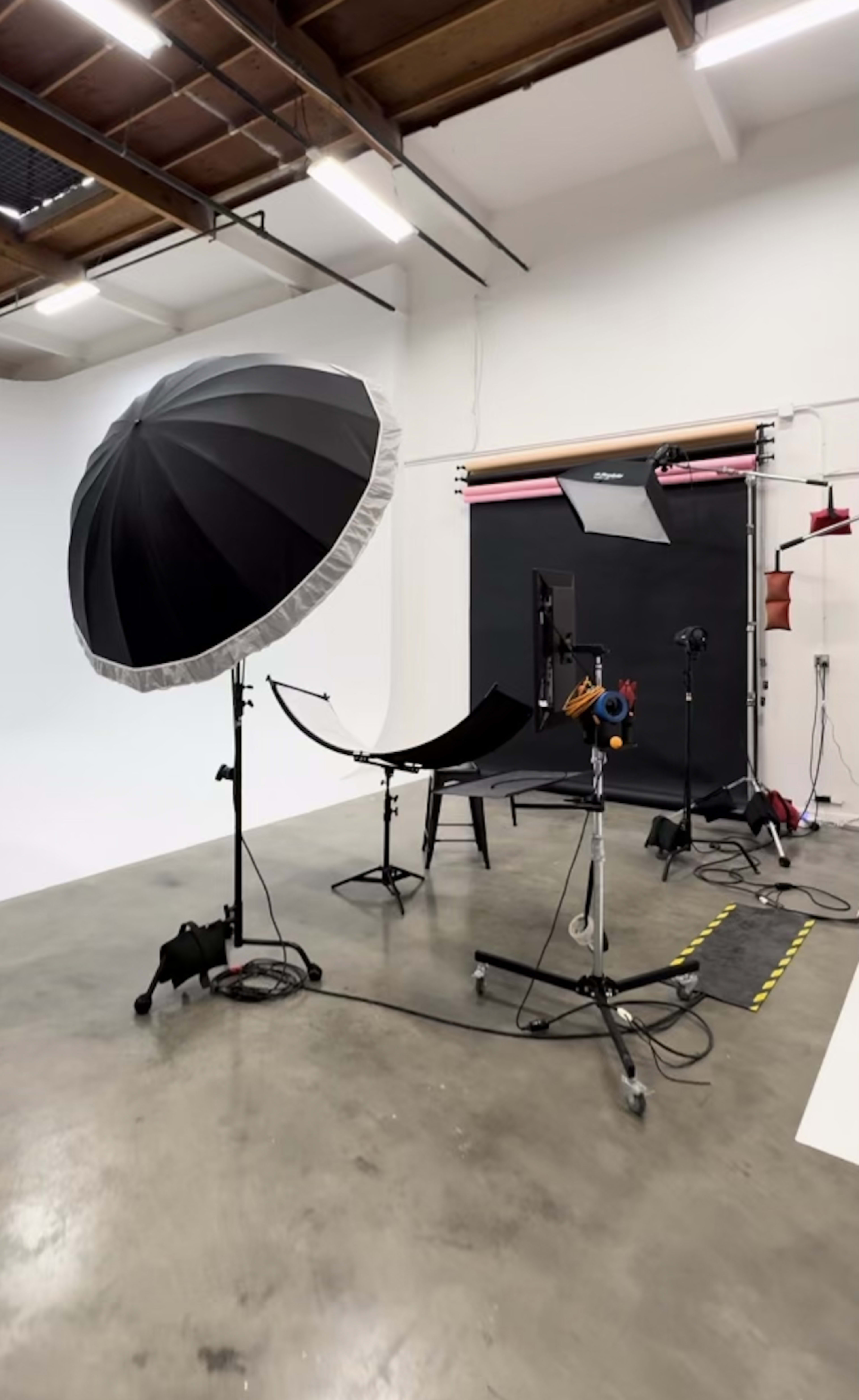 A photography studio setup featuring a large black umbrella light, a backdrop stand with a black curtain, and various equipment on the concrete floor.