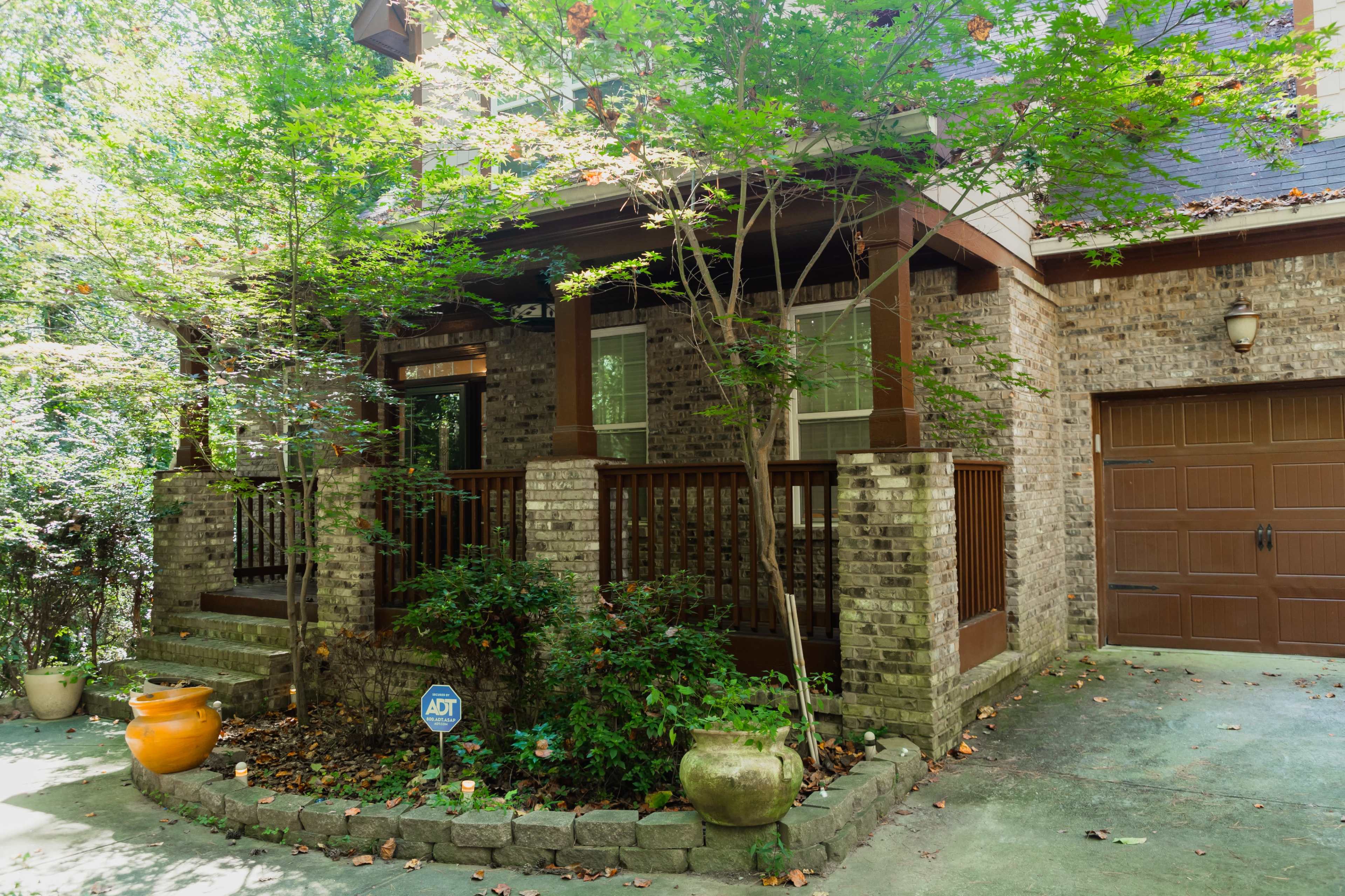 The image shows a brick house with a front porch surrounded by greenery and a garage door to the right.