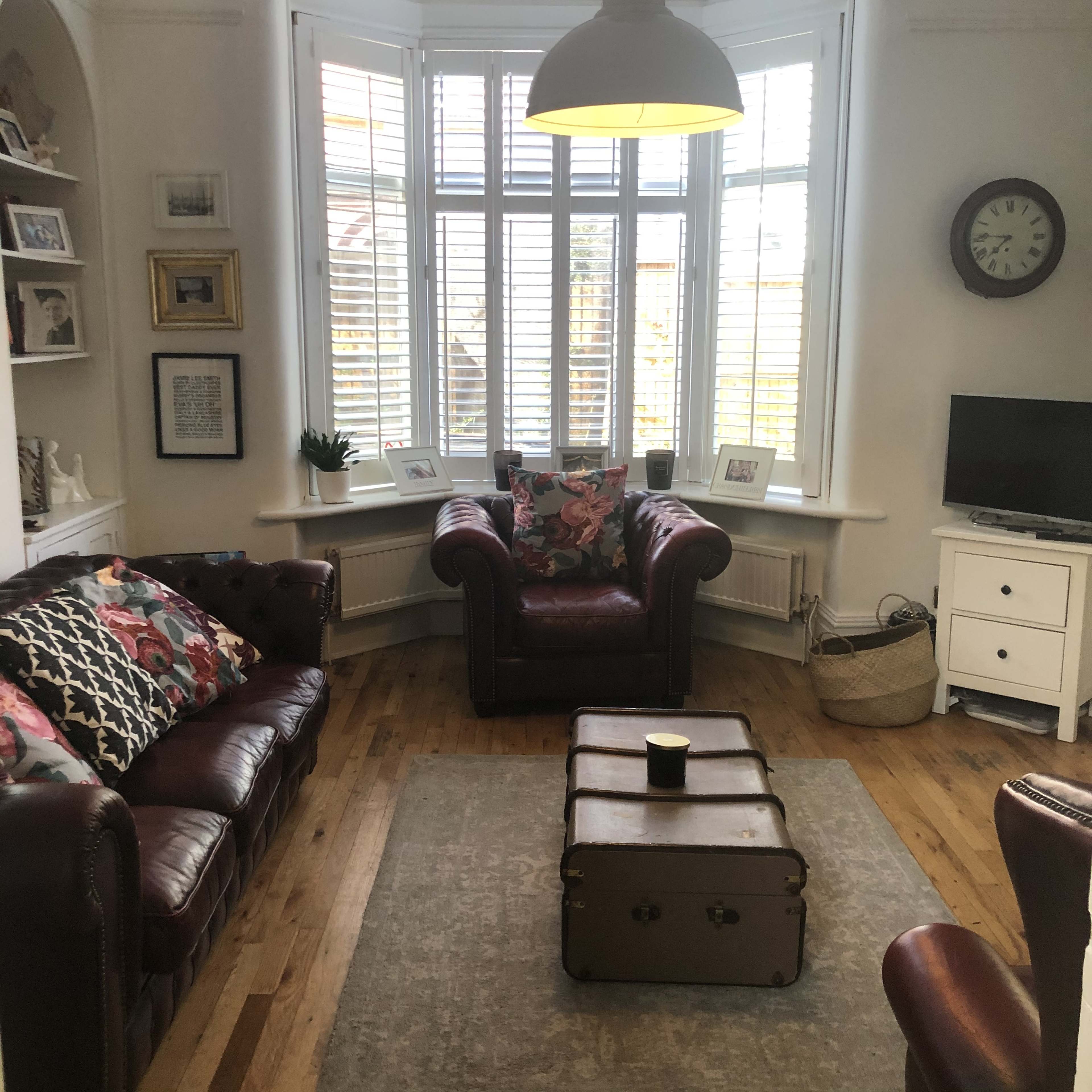 A cozy living room features two leather armchairs, a wooden coffee table, and a large bay window with shutters.