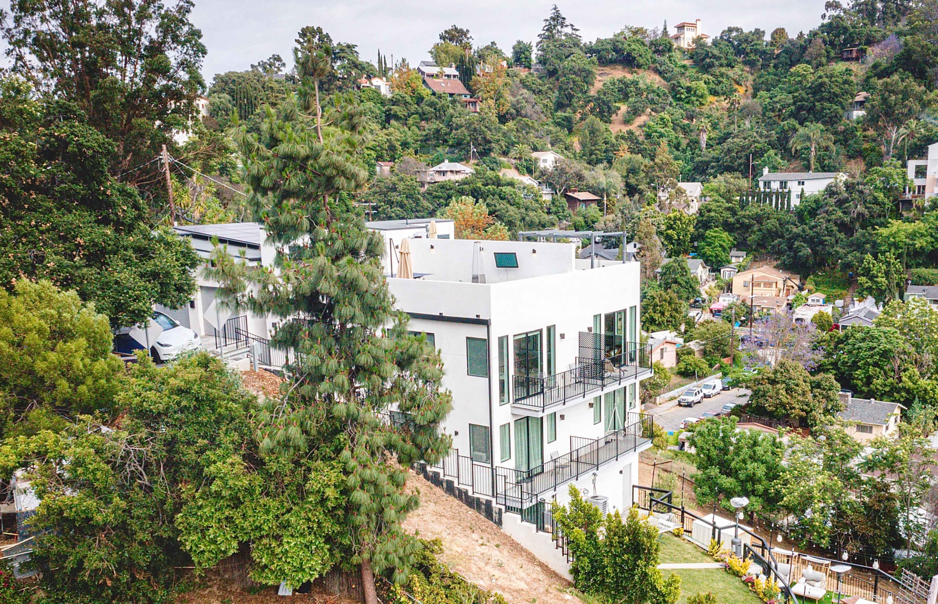 A modern white house with large windows and multiple balconies is situated on a hillside surrounded by greenery.